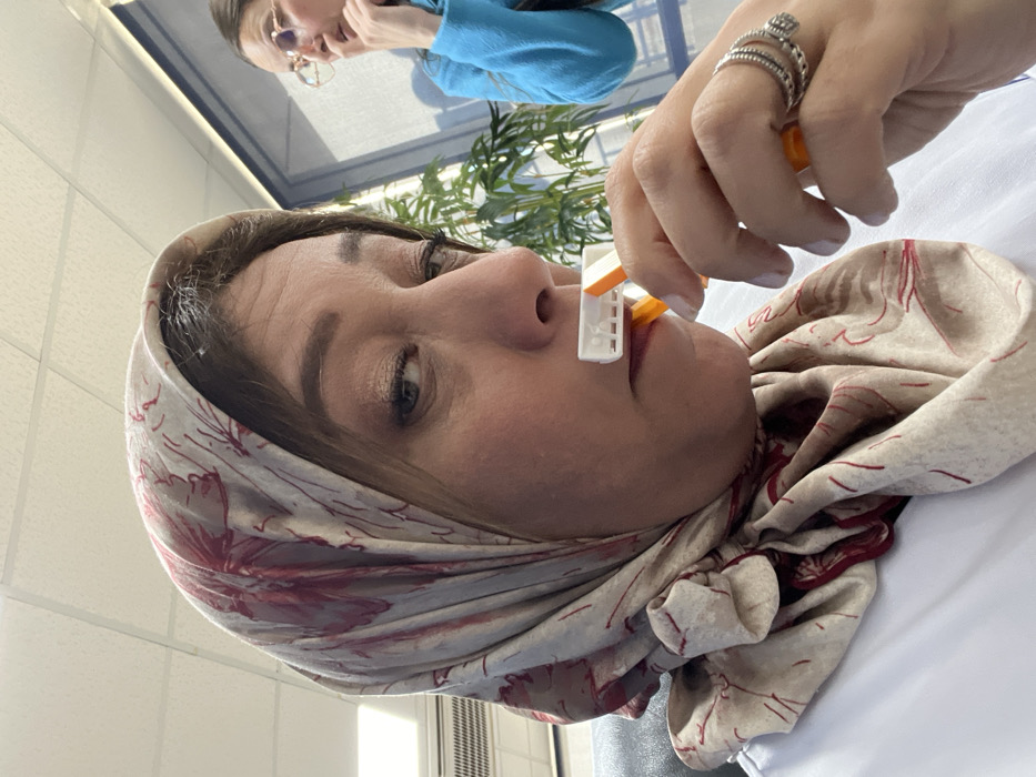 A woman wearing a floral headscarf has a device clipped to her nose, held by another person's hand adorned with rings. The device is white with orange accents. The woman looks slightly upward with a calm expression. In the background, a woman with glasses and a blue shirt is partially visible, along with a blurred green plant. The setting appears to be indoors, possibly in a medical or clinical environment, with white walls and fluorescent lighting overhead.
