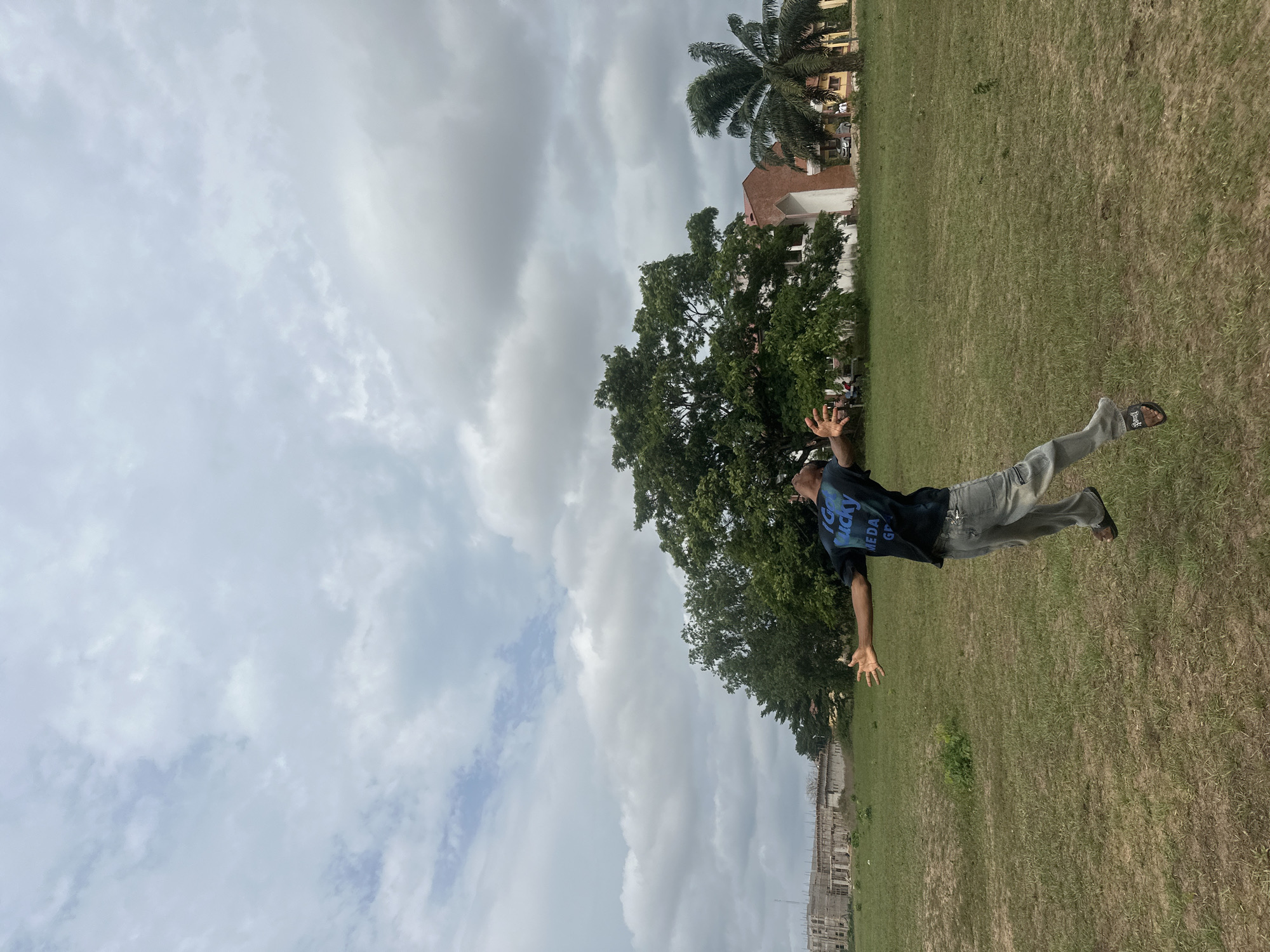 A male individual is captured mid-jump over a large, partially overgrown grassy field in Ihiagwa, Nigeria, under a heavily clouded sky. The subject, viewed from slightly behind and to the right, wears a dark t-shirt with blue text, light-colored jeans, and dark sandals, with both feet off the ground. His arms are outstretched laterally, palms open, and his head is tilted backward, looking upwards.
In the background, to the left, multiple multi-story concrete structures are under construction, with visible rebar and unfinished facades. Towards the center and right, a cluster of mature green trees, including a large deciduous tree and a prominent palm tree, partially obscure finished buildings. These buildings feature lighter colored walls and red or reddish-brown roof sections and facades. Cars are visible parked near these finished structures. The field foreground consists of mixed green grass and bare earth patches.