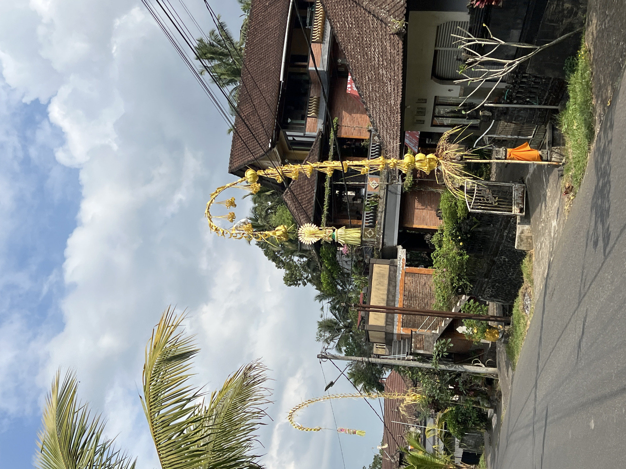 The image depicts a street scene in Pupuan, Indonesia, under a partly cloudy sky. On the right side of the asphalt road, there is a house with a dark brown tiled roof and yellow walls. A decorative, yellow, arched structure adorned with yellow spheres and foliage stands in front of a gate. An orange fabric is tied to a pole supporting this structure. To the left of the road, a second, similar yellow arched decoration is visible, hanging from a pole and also featuring yellow spheres and foliage. Further down the road, more houses with brown tiled roofs and brick accents are visible, surrounded by lush green vegetation and palm trees. Electrical wires crisscross overhead. The scene appears calm and evokes a sense of daily life in a tropical environment.