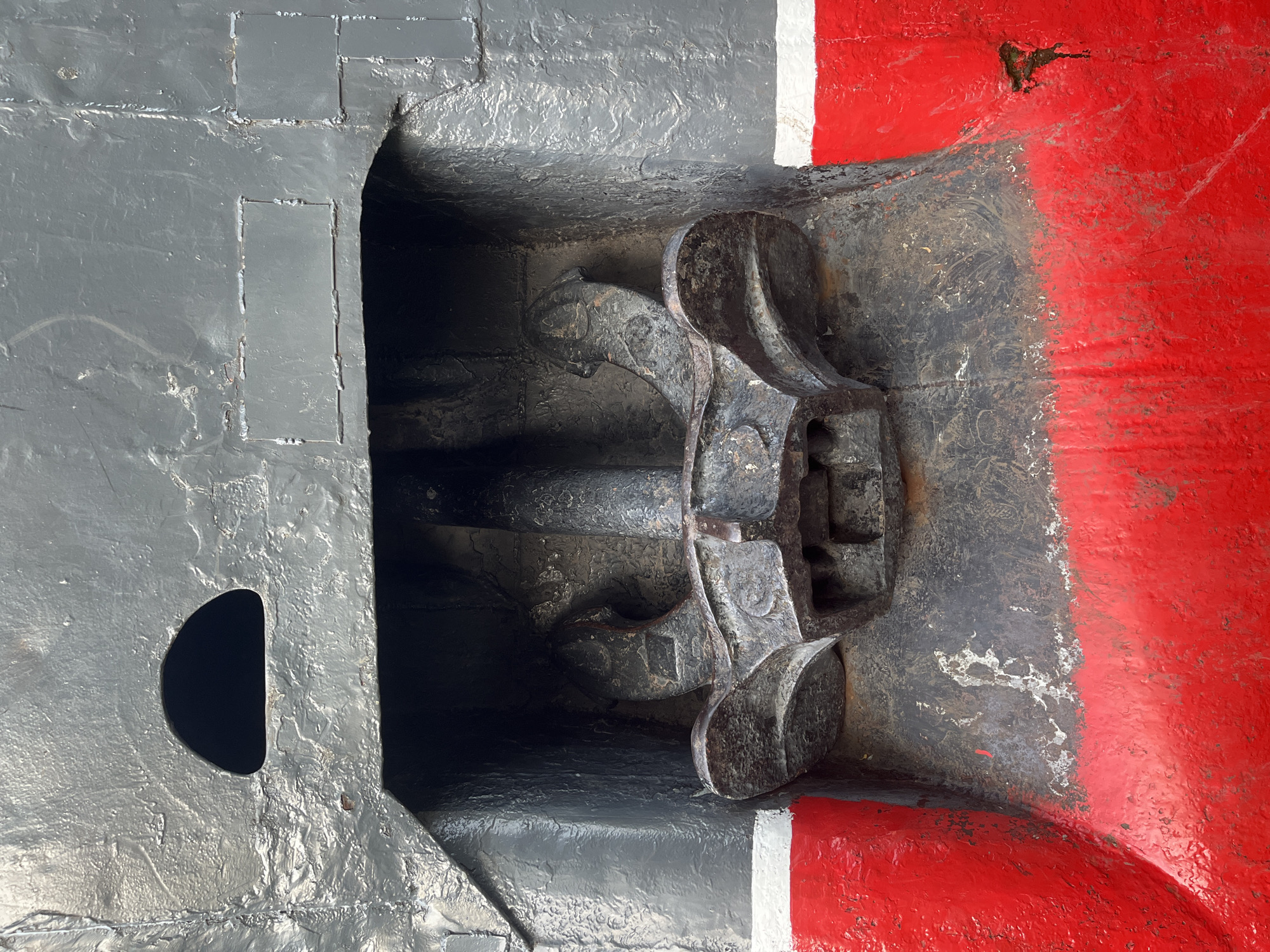The image shows a metal anchor stowed within a recess in the bow of a ship. The ship's hull is painted in shades of gray and red, separated by a white stripe. The anchor is dark and appears weathered. A semicircular opening is visible on the gray portion of the hull near the anchor recess. This image appears to be from the Krasnodar region of Russia.