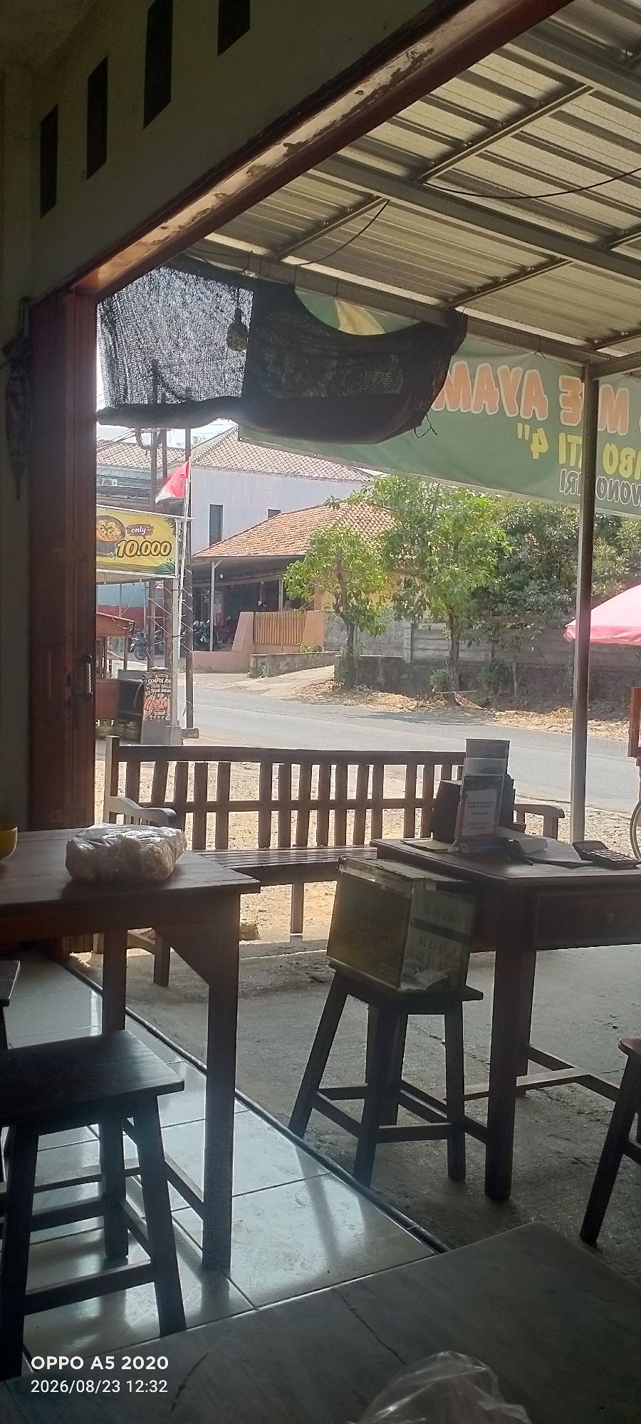 The image captures an outdoor seating area of a casual eatery in Dusun Satu, Indonesia. Wooden tables and chairs are arranged on a tiled floor. In the foreground, a table holds a wrapped loaf of bread. Behind it, a wooden bench with a slatted back faces a street. A sign advertising "MAYA M" in green lettering is visible in the background, along with a red and white Indonesian flag. The environment suggests a warm, possibly sunny day, with trees and buildings visible beyond the seating area. The timestamp on the image indicates it was taken on August 23, 2026, at 12:32 PM, and was captured by an OPPO A5 2020 device.