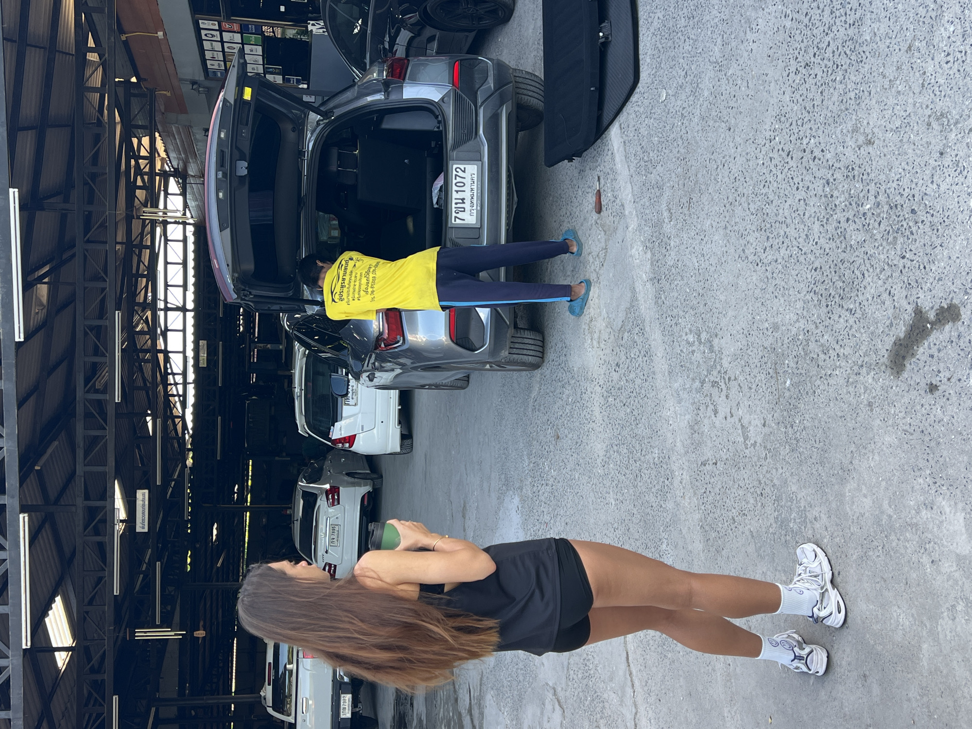 The image shows two women in a partially covered location likely a garage, possibly in Baan Lipa Noi, Thailand, given the license plates of nearby vehicles. One woman, dressed in a black top and shorts, holds a green container and is standing perpendicular to the camera, facing a row of cars. Another woman is working inside the open trunk of a gray car, wearing a yellow shirt and blue pants, she appears to be fixing something. The trunk of the car is open and a black cover is nearby. There are several other cars parked in the background, and a metal structure covers the parking area.