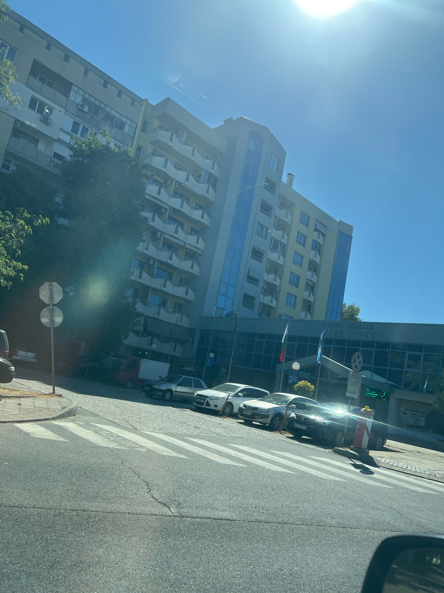 The image shows a street scene in Plovdiv, Bulgaria, on a sunny day. A large, modern building with a glass facade and balconies dominates the background. In front of the building, several cars are parked along the street, including a white Ford Focus, a silver sedan, and a black sedan. A pedestrian crossing is visible in the foreground, with white stripes painted on the asphalt. The sky is clear blue, with the sun shining brightly and creating lens flare in the upper right corner of the image. The overall impression is one of an urban environment bathed in bright sunlight.