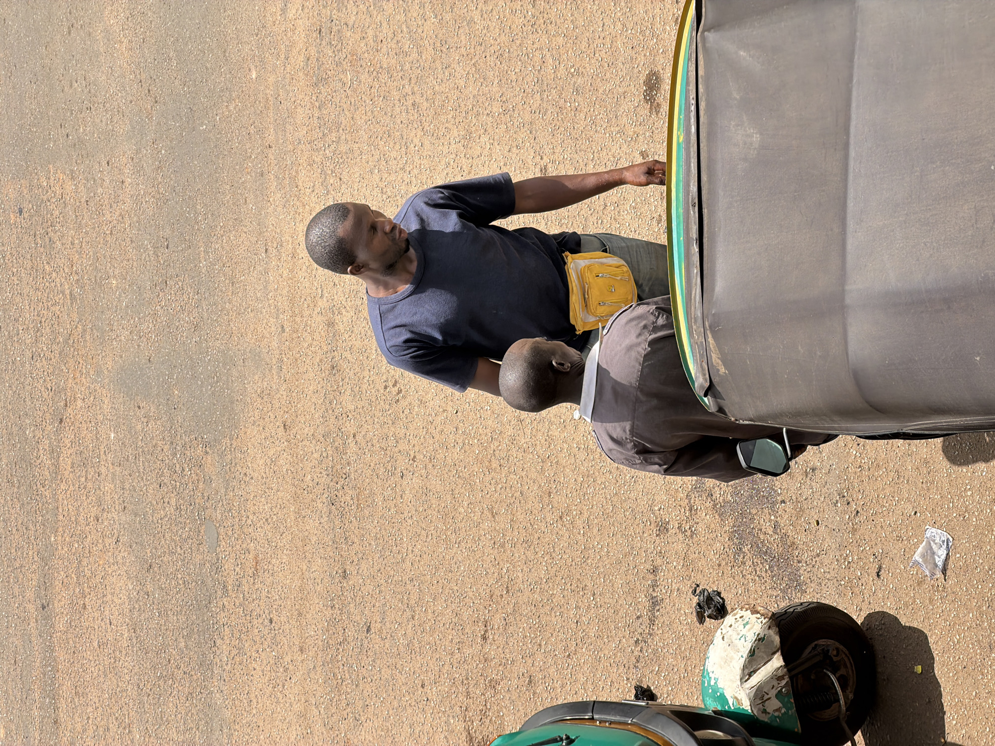 This image shows two men and a portion of a three-wheeled vehicle, likely a tuk-tuk, in Bukuru, Nigeria. The scene appears to be outdoors on a paved surface, possibly a street or roadside, with loose gravel and sand. The lighting suggests it is daytime, with bright sunlight casting shadows.
One man, wearing a dark blue t-shirt and jeans, stands and looks towards the vehicle. He has a yellow fanny pack around his waist. The second man is partially visible, bent over or seated, and wearing a brown shirt and grey pants. His head is towards the foreground, and his body is mostly obscured by the vehicle.
The three-wheeled vehicle is predominantly grey and green. A wheel and part of the chassis are visible in the foreground. The overall impression is a casual, everyday scene, possibly a stop on a journey or a moment of rest. There is no visible text in the image.