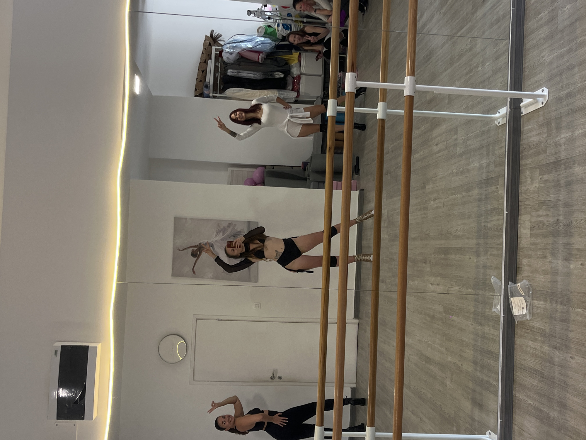 A mirrored wall in a room reflects images of women and furniture. The mirrored wall includes ballet barres, white baseboards, and a slim light strip to the left. Reflected objects include: at the bottom of the frame, a woman in black athletic clothes posing; in the middle, a woman in black athletic clothing taking a selfie; further back, a woman in a white dress is posing with her hand raised; in the background, clothing hanging on a rack and two women. A door and a ballet painting are visible in the reflected image, along with a small, round mirror. A package is attached to the mirror in the foreground. The floor has a light gray woodgrain pattern. The location is possibly a dance studio in Budva, Montenegro.