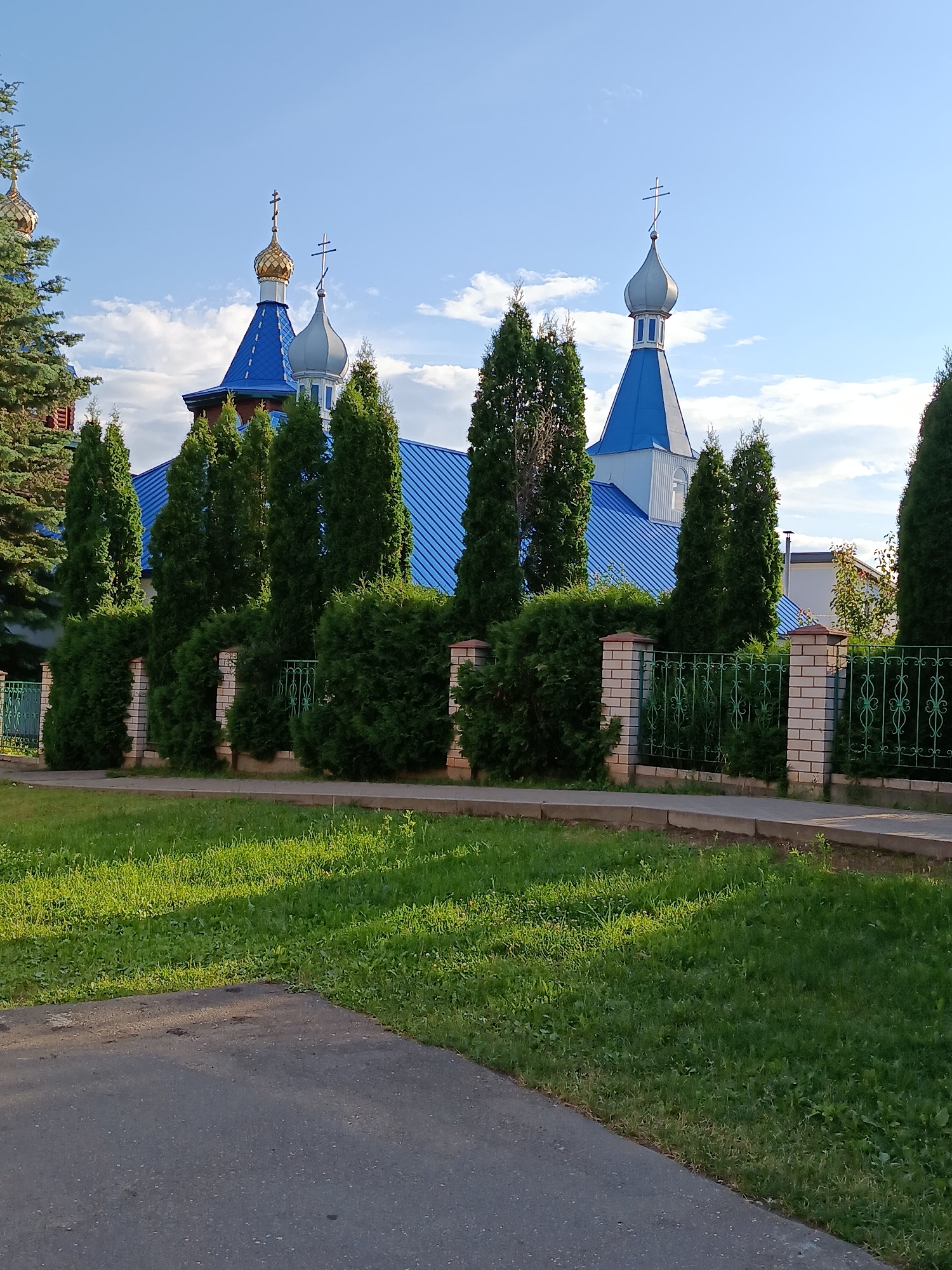 The image shows a view of a church with blue roofs and golden and silver domes, topped with crosses, under a bright blue sky with scattered clouds. A fence made of brick pillars and green metal bars runs along the foreground, with a well-maintained hedge and several tall evergreen trees behind it. To the left of the church, a large evergreen tree is visible. In the foreground, a patch of green grass is illuminated by sunlight, with a paved area in the lower left corner. The scene appears to be a public space, possibly a park or the grounds of the church, located in the vicinity of Смілавіцкі сельскі Савет, Belarus. No people are present in the image.