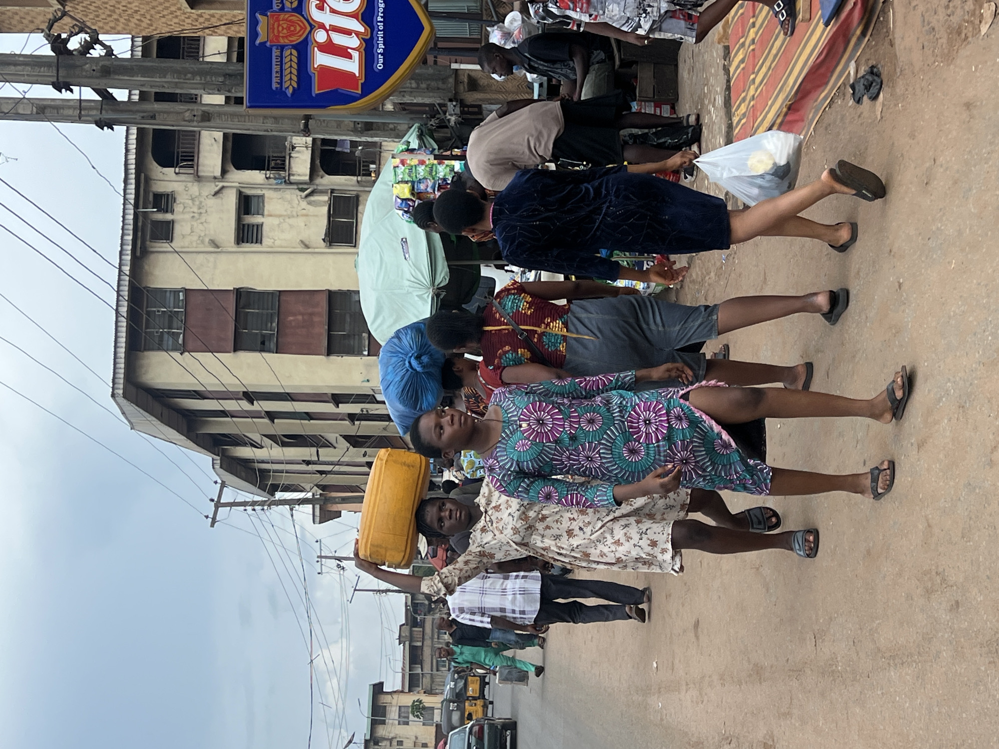 In Nkpor, Nigeria, a street scene unfolds under an overcast sky. A woman in a floral dress balances a bright yellow jerrycan on her head as she walks forward. Beside her, a young woman in a patterned green and purple dress walks in the same direction, her head down. Behind them, other individuals are also moving along the dirt and asphalt road, some carrying bundles on their heads. The background features multi-story buildings with weathered facades and balconies. Electrical poles and wires crisscross the sky above. A street stall with a green awning displays various items, and a blue and yellow sign for "Life" beverage is visible on a pole. The scene captures the daily life and movement of people in a busy Nigerian urban environment.