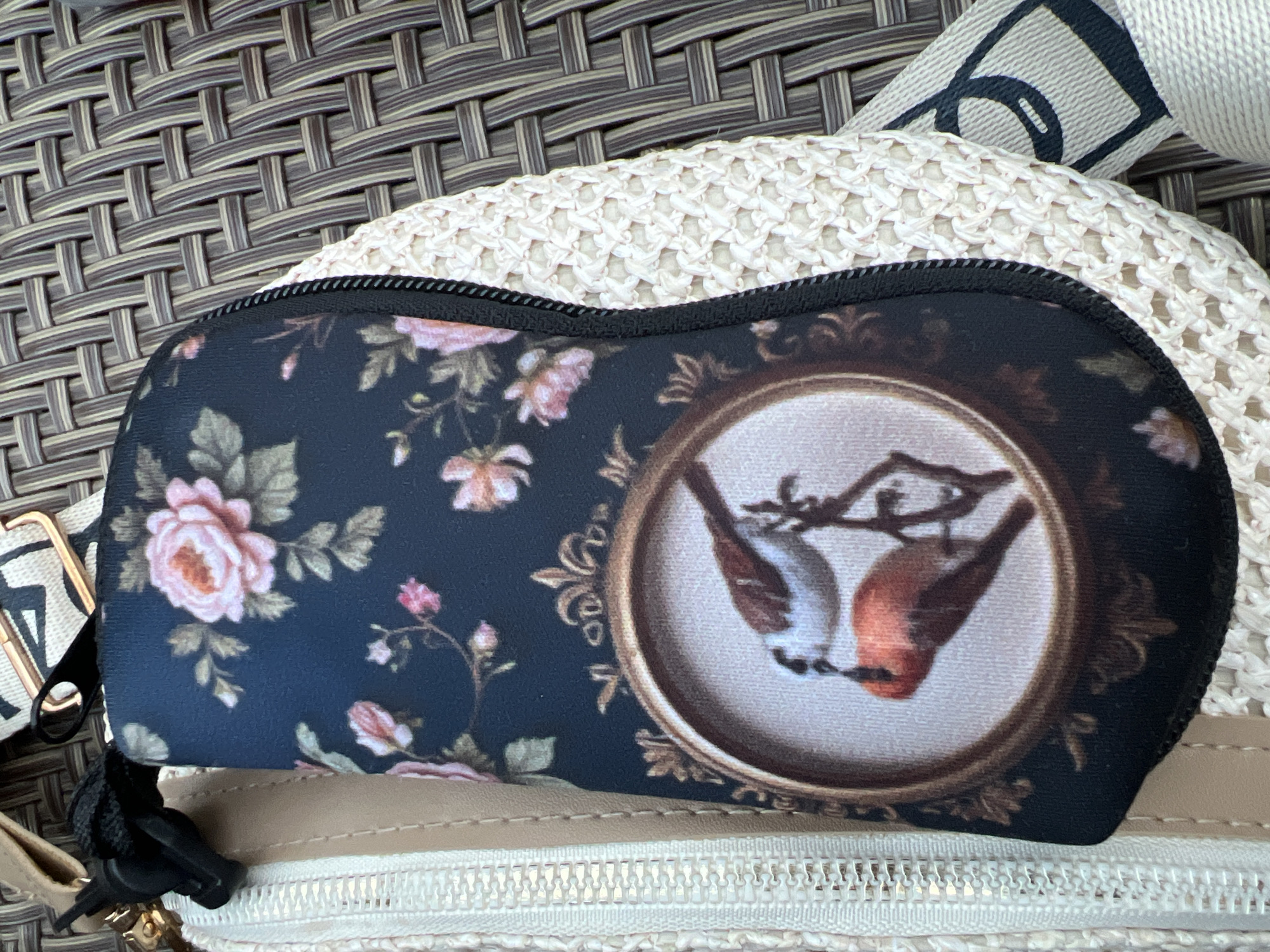 A close-up, overhead shot shows a black zippered pouch with a floral pattern and a circular vignette of two birds on a branch. The pouch is resting on a light-colored, crocheted surface, which is on top of a woven brown material. A tan strap with a black and white pattern is partially visible, along with metal hardware and a black lanyard clip. The scene is outdoors, as indicated by the textured surface that resembles outdoor furniture. There are no people or animals present in the image, nor any identifiable landmarks that would place it in Bender, Moldova.