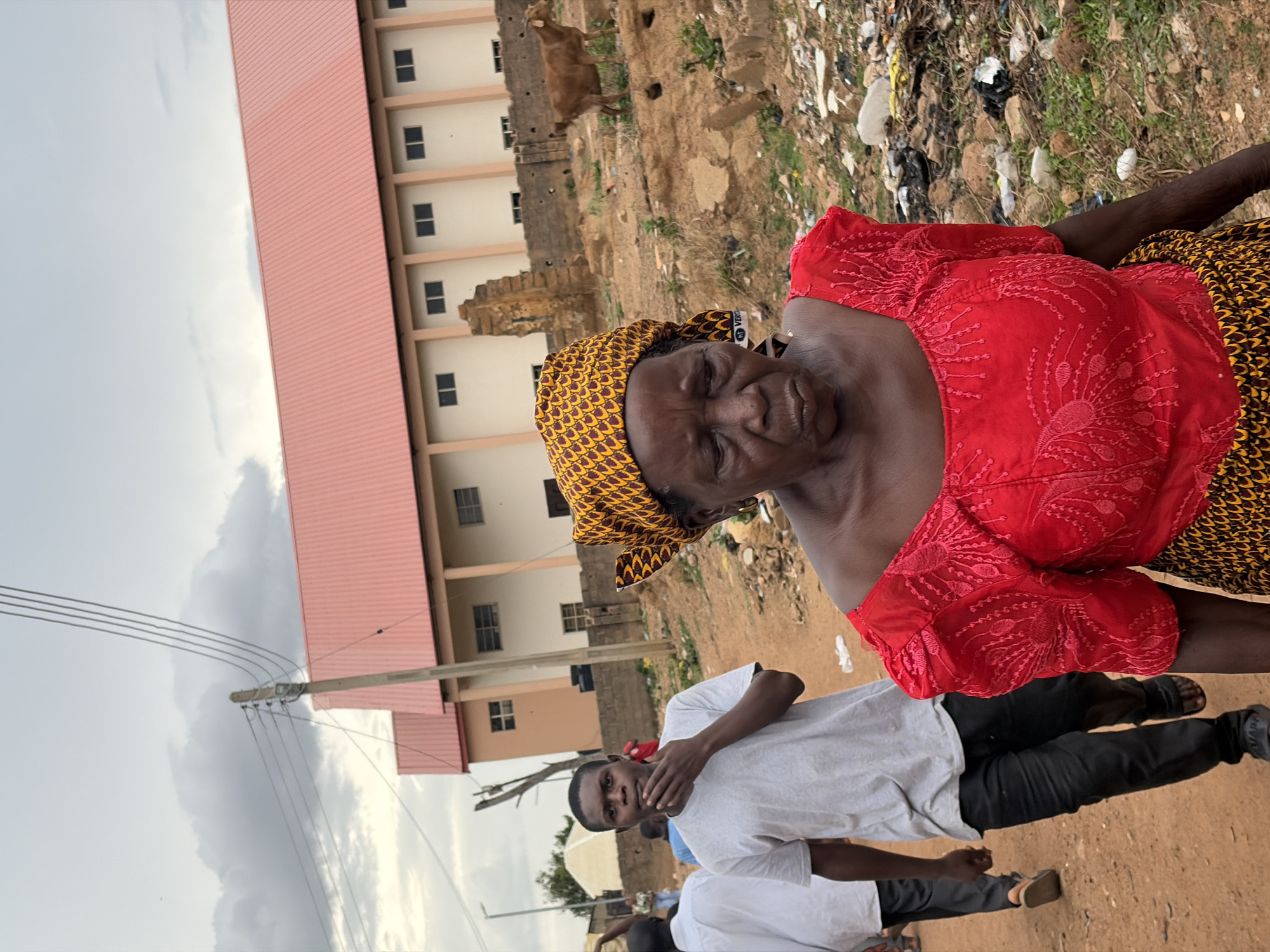 An image of an older woman wearing a red top and patterned head covering stands in front of a multi-story building with a red roof in what appears to be Jos, Nigeria. A young man in a white t-shirt is visible in the foreground. A goat is present in the background near a trash pile. Overhead power lines are also present. The scene suggests an urban setting with evidence of environmental issues.