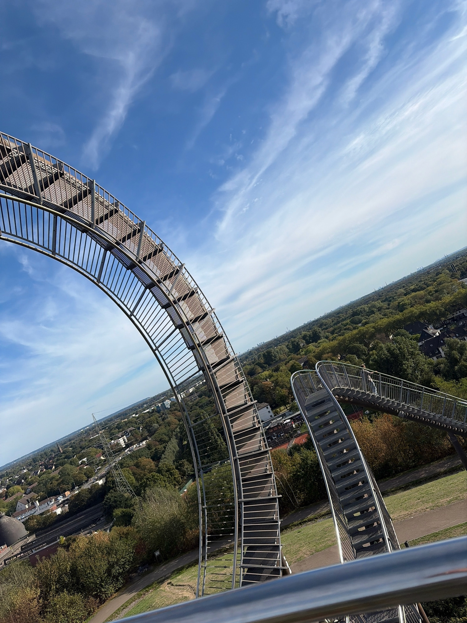 The image captures a high-angle view from the "Tiger and Turtle – Magic Mountain" sculpture in Duisburg, Germany. The metallic structure, featuring a spiral staircase and walkways, dominates the foreground, curving upwards against a clear blue sky dotted with wispy white clouds. Below the sculpture, the cityscape of Duisburg unfolds, characterized by a dense canopy of green trees, interspersed with residential buildings and industrial elements like a large pylon. The overall impression is one of an urban landscape viewed from an elevated artistic installation on a bright, sunny day.