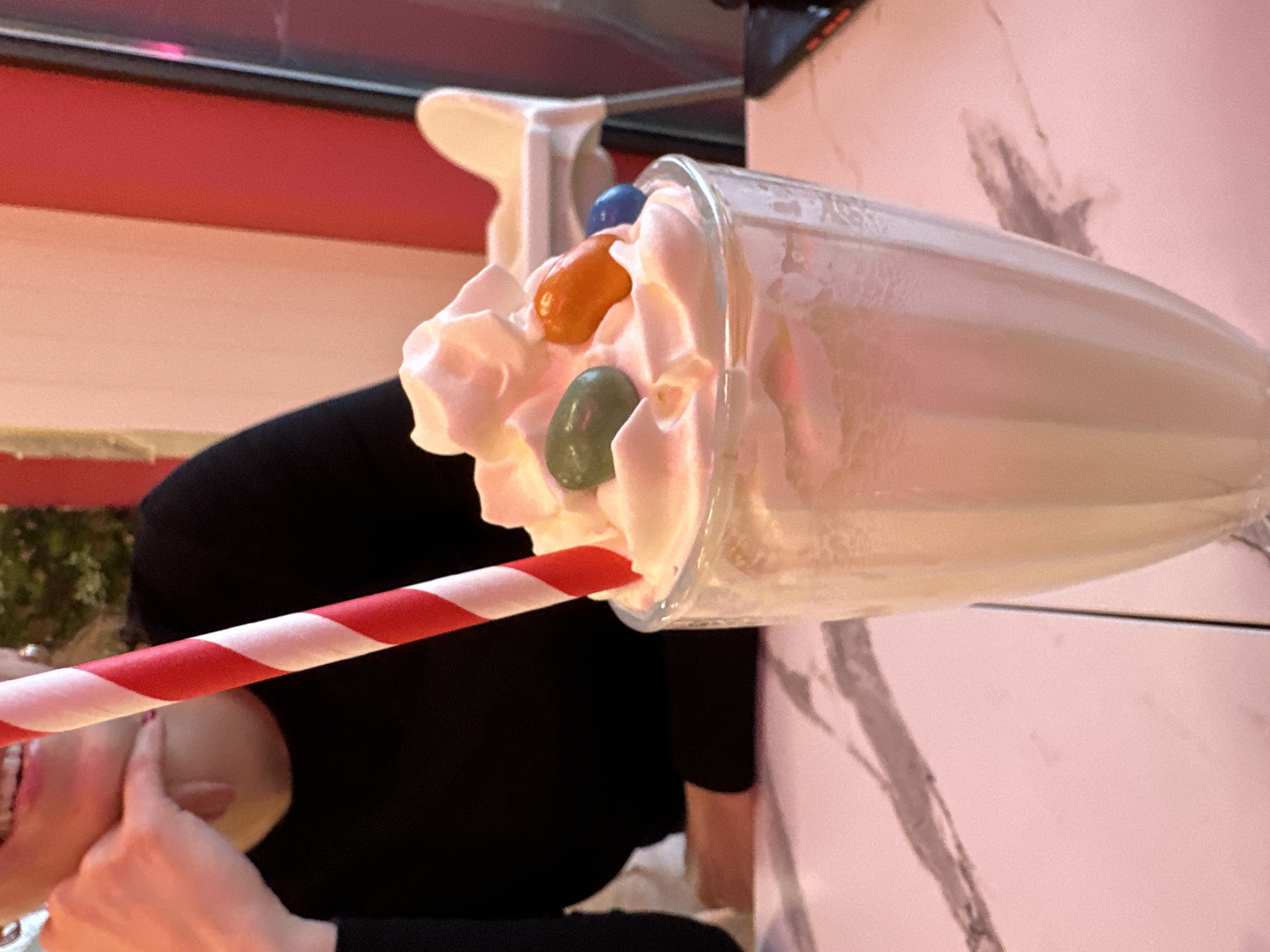 A photograph taken in Sofia, Bulgaria, presents a stylized milkshake. The milkshake, topped with whipped cream and colorful candies, sits in a tall, clear glass. A red and white striped straw is inserted into the drink. The milkshake is positioned on a surface with a marble-like pattern. The background features a person wearing a black garment.