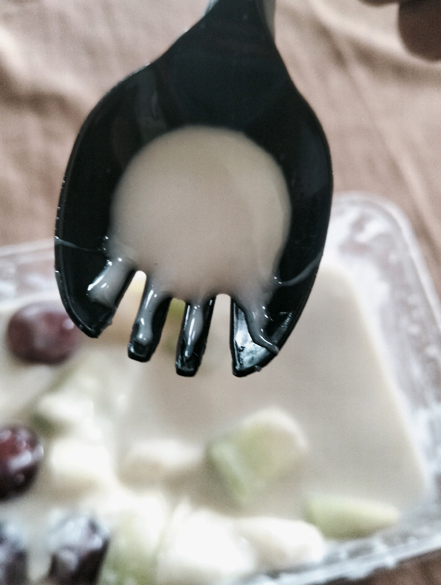 A close-up, overhead shot shows a black plastic fork lifting a dollop of creamy white pudding, possibly yogurt or custard, from a clear plastic container of fruit salad. The pudding drips from the tines of the fork. The container, out of focus in the background, holds chunks of what appear to be green melon, white fruit, and dark red grapes. The scene is set on a textured, light brown surface, likely a tablecloth or mat. No specific location in the city of Medan, Indonesia, can be determined from this image.