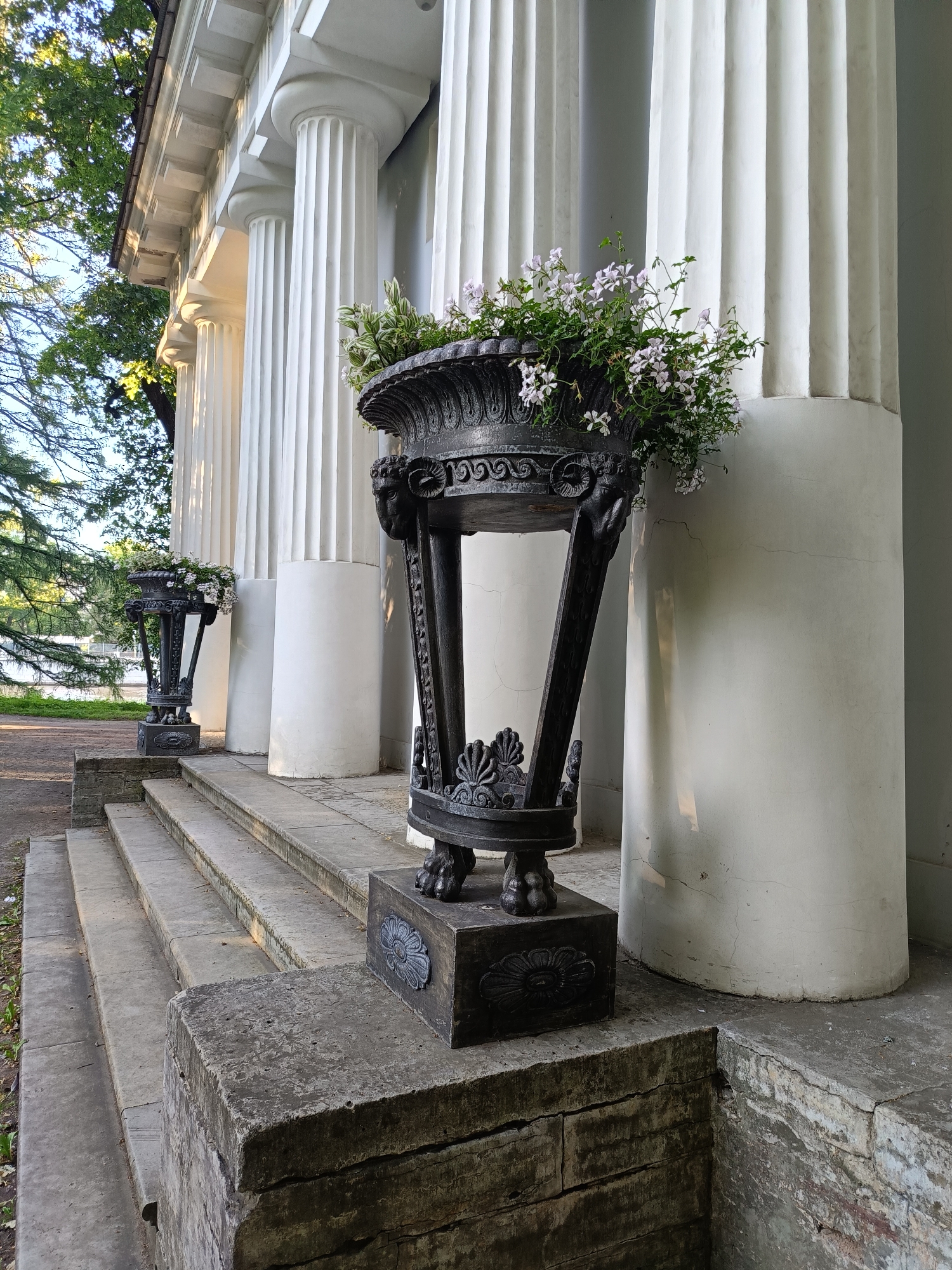 Two ornate black metal planters, each filled with light pink flowers and greenery, are placed on either side of a set of stone steps leading up to a white building with large fluted columns. The planter in the foreground is larger and more detailed, featuring ram's head decorations. The planter in the background is smaller and simpler. The steps are made of weathered stone and are lined with stone blocks. The building appears to be an old, classical structure with a light-colored facade. Trees with green foliage are visible behind the building and to the left. The lighting suggests it is daytime, with sunlight casting shadows on the columns and steps.