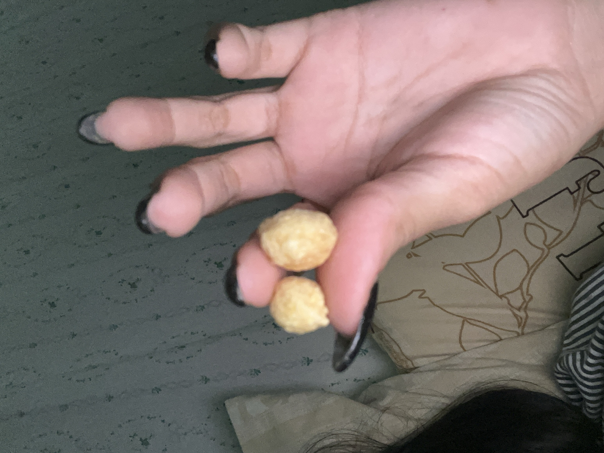 A close-up shot focuses on a person's hand holding two small, round, light-yellow objects that appear to be cereal or similarly processed food items. The hand has long, dark nails. The background includes a bed with patterned pillows and sheets, as well as a glimpse of the person's hair. The location cannot be determined to be Santa Ana, Venezuela from the content of the picture.