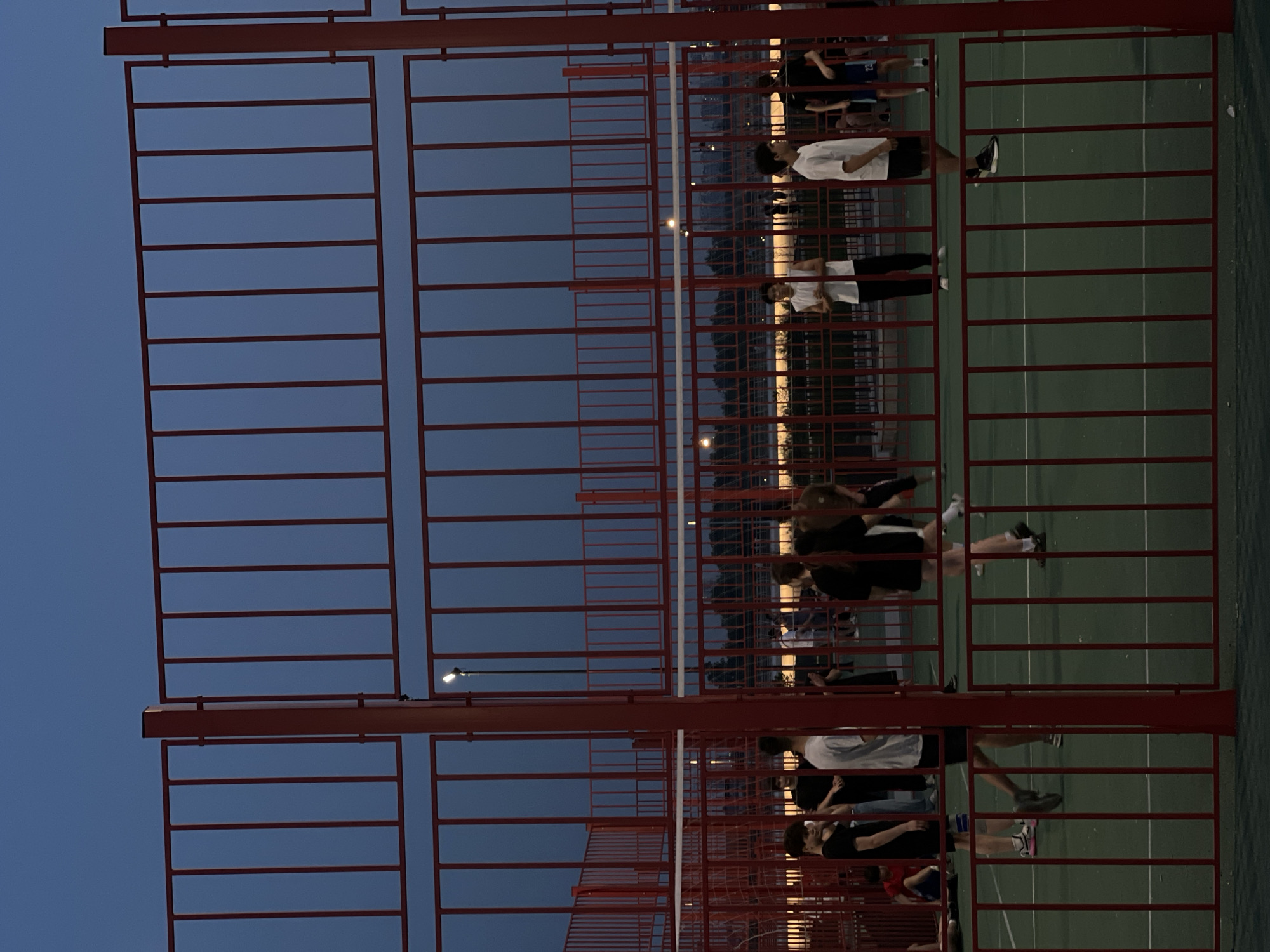 A ground-level, medium-range shot shows a fenced-in outdoor recreational area with multiple people on the green playing surface. The location is identified by the fencing style as being in Moscow, Russia. A red metal fence, composed of horizontal and vertical bars, dominates the foreground. Various people are visible within the fenced area, engaged in activities like standing, walking, or interacting with others. The lighting appears to be twilight or early evening. The sky visible above the fence has a gradient of blue hues. Streetlights are seen in the background, suggesting an urban environment. The ground within the fenced area is green and appears to be synthetic turf.
