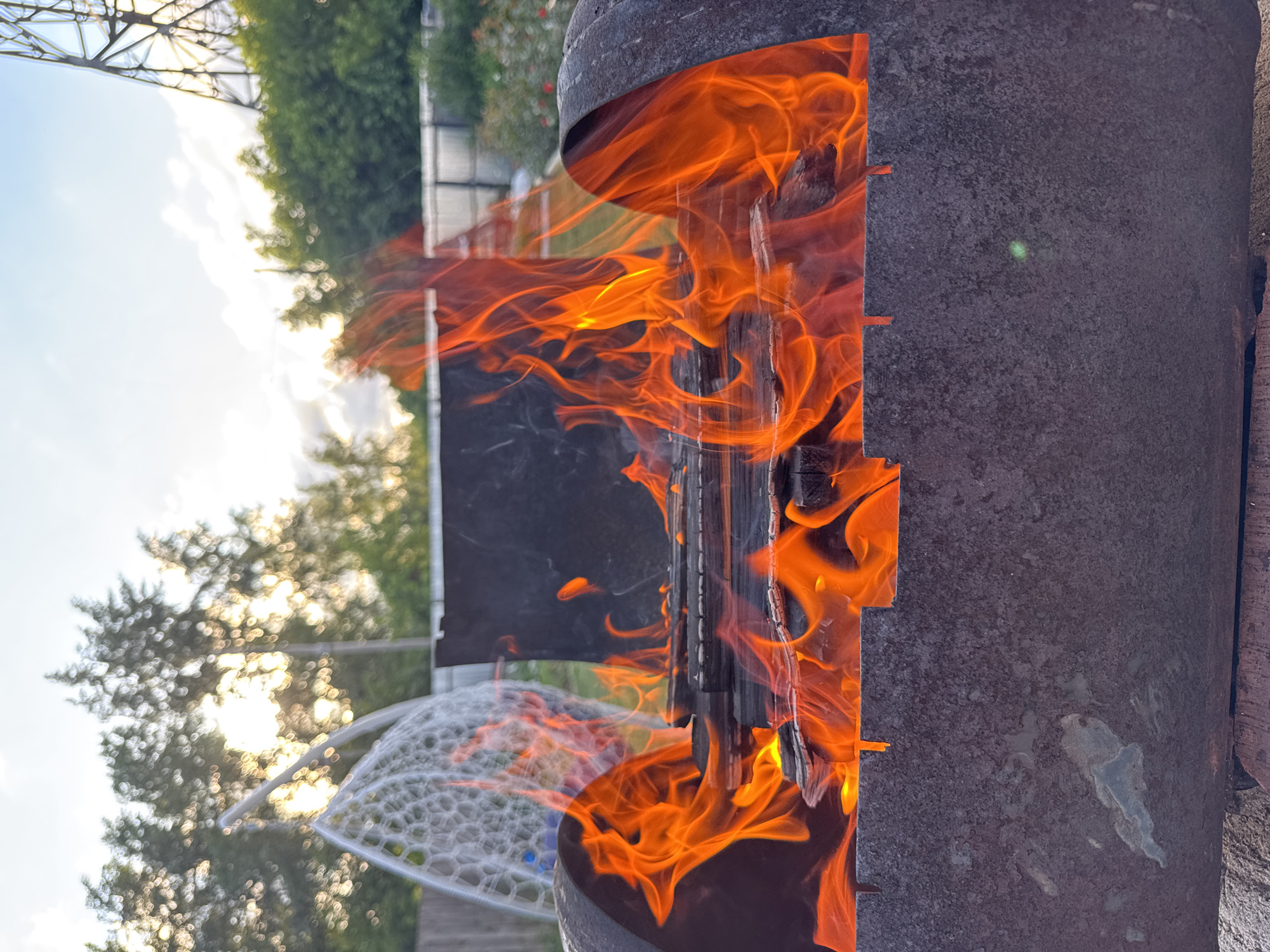 The image depicts a close-up view of a fire burning in a metal brazier. The fire is the dominant element, with vibrant orange and yellow flames consuming the wood. The brazier appears to be made of weathered metal, with visible rust and texture. In the background, a soft-focus scene of trees and sky is visible, suggesting an outdoor setting. The lighting indicates it is likely late afternoon or early evening, with the sun's rays filtering through the trees. There are no people or animals clearly visible in the frame. The environment suggests a casual outdoor gathering or a moment of relaxation in a backyard or park area. The presence of a swing-like structure to the left and a tall metallic tower in the far background hint at a residential or recreational area, possibly on the outskirts of a town or city. The overall impression is one of warmth and natural ambiance. The city of Chaikovsky, Russia, is a town located in the Perm Krai, known for its industrial enterprises and proximity to the Kama River, a major waterway in European Russia.