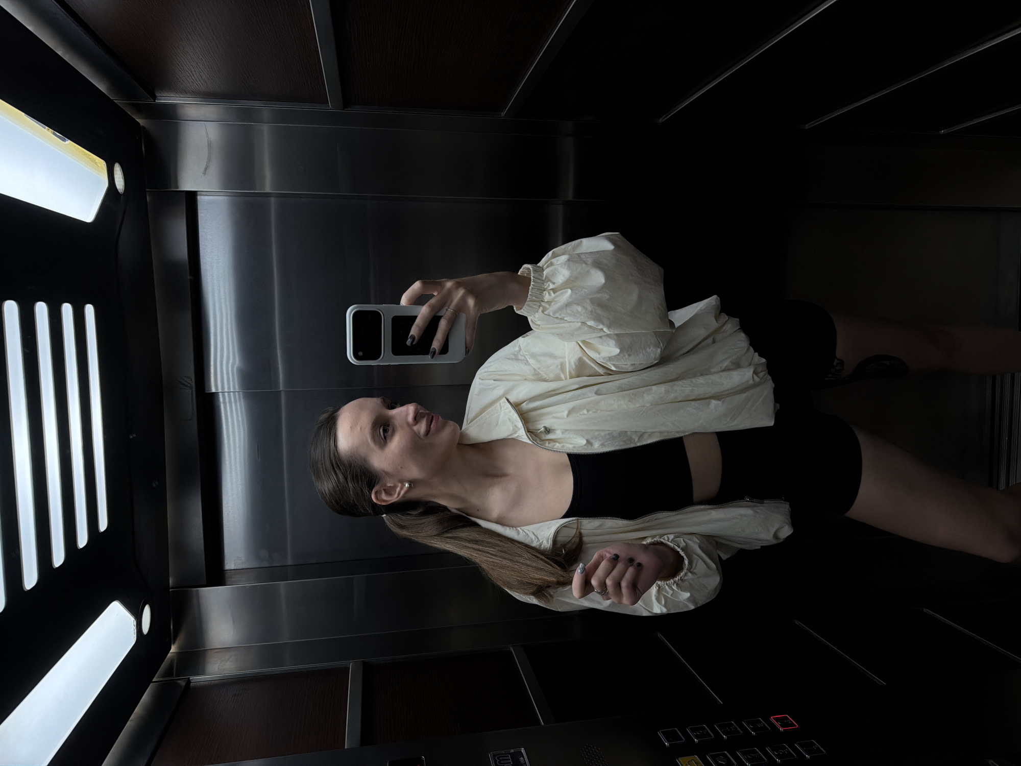 A woman is taking a selfie in an elevator. She is wearing a black crop top, black shorts, and a white jacket. The elevator has stainless steel walls, panel lighting, and control buttons. The location is inside an elevator in Budva, Montenegro, based on visual clues.