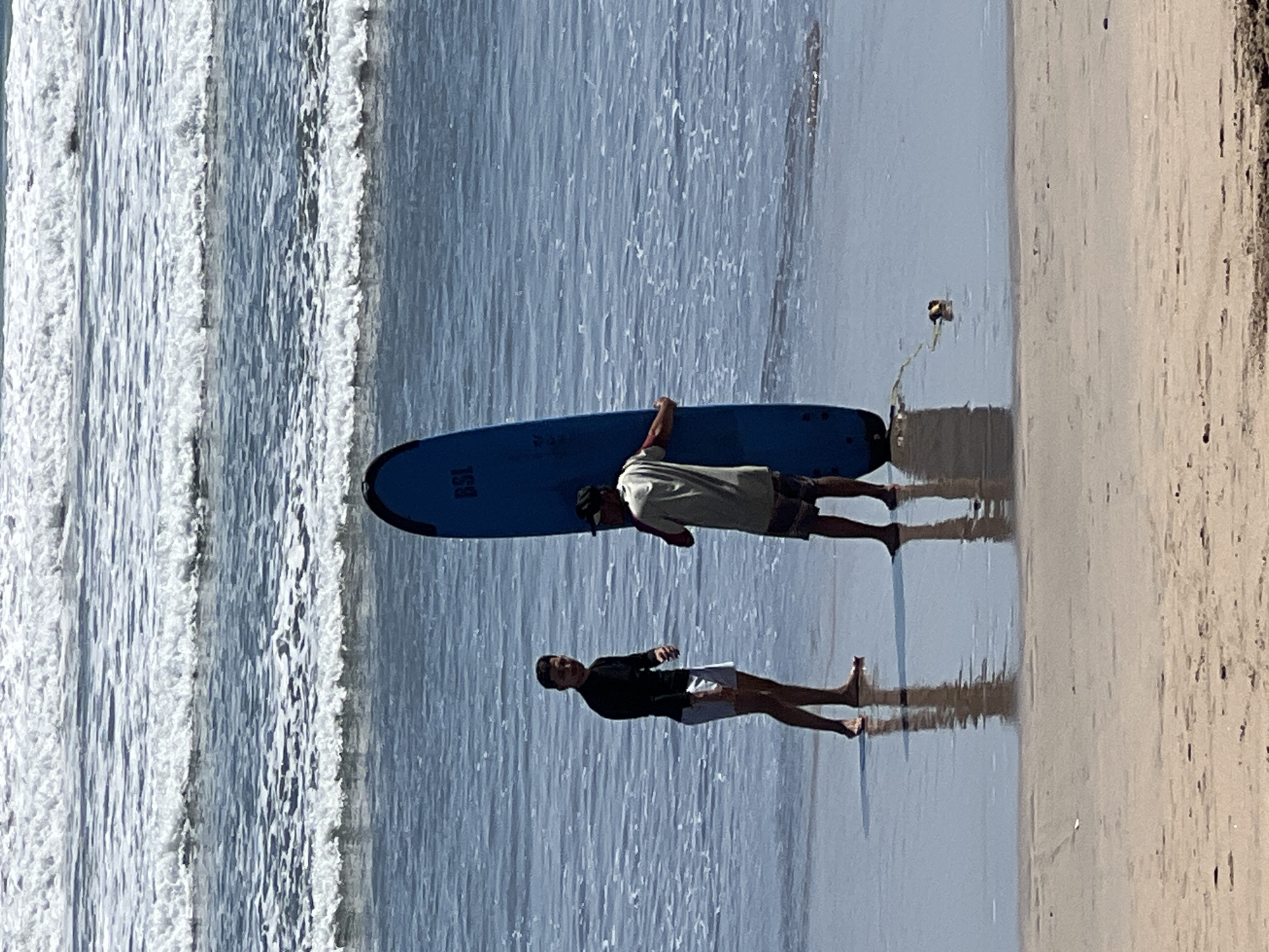 Two individuals stand on a wet sandy beach in Kuta, Indonesia, with the ocean and waves in the background. One person, on the left, is wearing a black long-sleeved shirt and white shorts, facing the camera with their left hand extended. The second person, on the right, is wearing a light-colored t-shirt and patterned shorts, holding a large blue surfboard upright. The surfboard has text that reads "BSL." A yellow leash is attached to the surfboard and trails into the water. The water shows gentle waves and ripples. The sand is wet and reflects the figures and the sky.