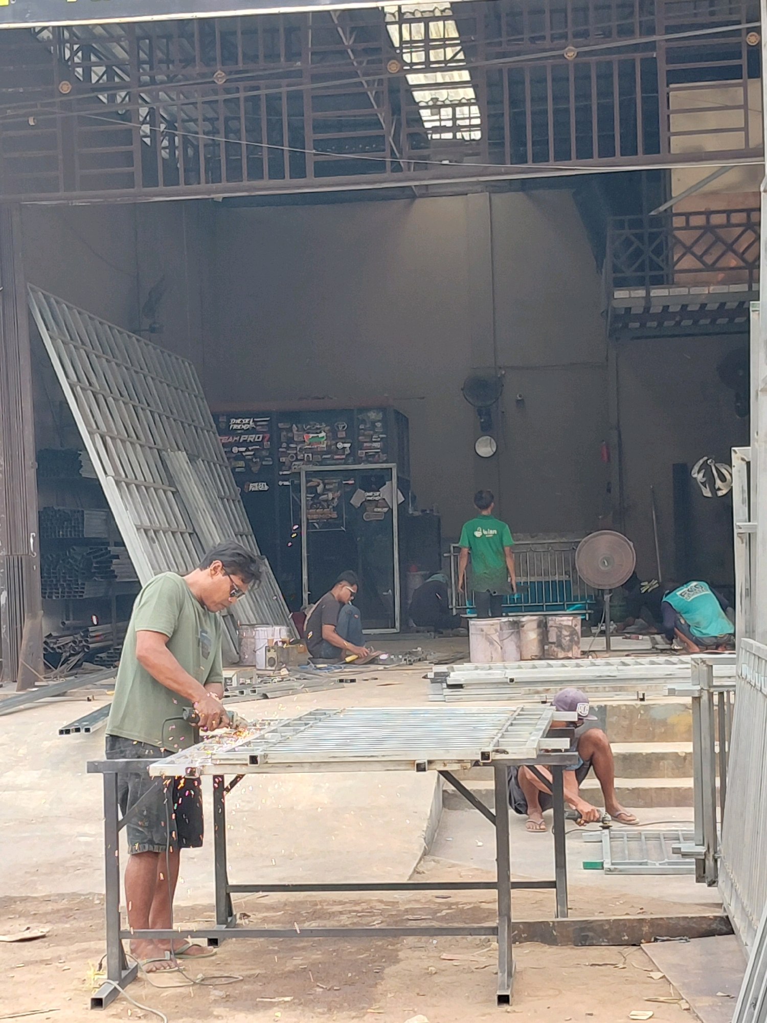 In the city of Persiapan Mulya Raya, Indonesia, a metal fabrication workshop is captured. The scene is dominated by raw materials and ongoing construction work. A man in a green t-shirt and shorts is actively grinding metal on a workbench, with sparks flying around him. To his right, another man is also engaged in metalwork, sitting on the ground and using a tool. In the background, several other individuals appear to be working, with one man in a green t-shirt standing and others either sitting or working on materials. Various metal structures, including large sheets and frames, are stacked and placed around the open-air workspace, suggesting a production or assembly line. The environment is industrial, with a dirt ground and a covered structure overhead supported by metal beams.