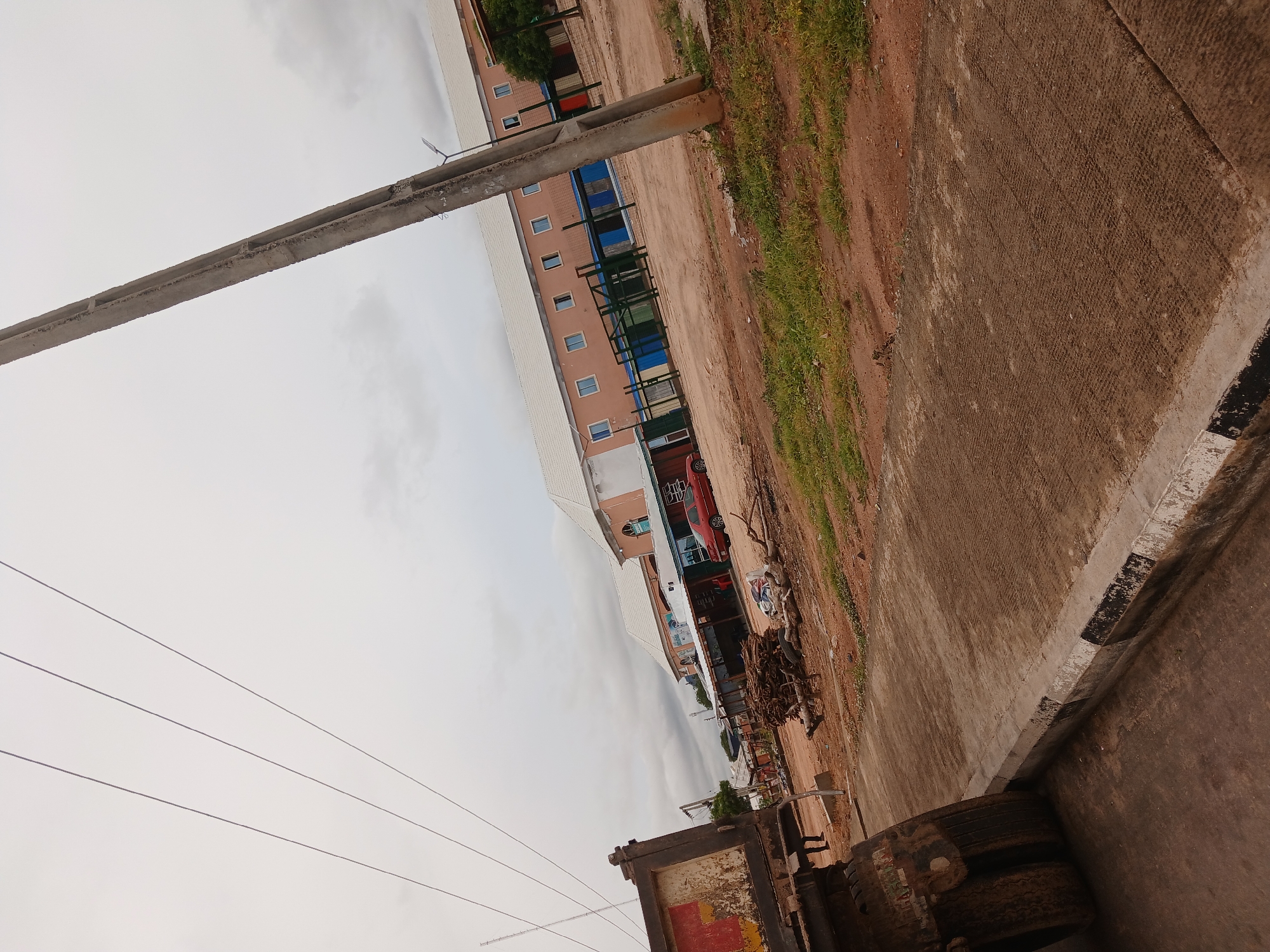 The image shows a dusty, partly paved area in Jalingo, Nigeria. In the foreground on the left is the rear of a large, weathered truck, its large tires and weathered metal body visible. A concrete curb with black and white markings separates the road from a patch of grass and dirt. In the middle ground, a pile of what appears to be firewood or branches sits on the dirt. Behind this, a red sedan is parked in front of a row of shops. The shops have various signs and displays, some with blue and green fencing. Further in the background, a long, light-colored building with multiple windows and a white roof dominates the scene, with more buildings and structures to its right. A tall concrete utility pole stands prominently in the right mid-ground. The sky is overcast and cloudy.