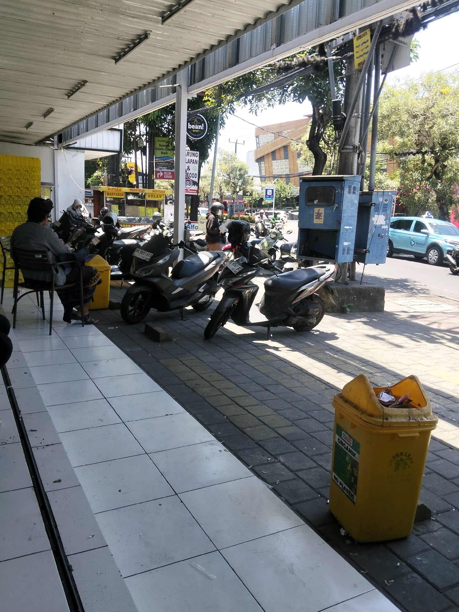 The image captures a street scene in Kuta, Indonesia, likely near a commercial establishment. A man in a gray long-sleeved shirt and dark pants sits on a black metal chair on a tiled walkway. In front of him is a yellow trash bin. To his right, several motorcycles are parked along the sidewalk. Beyond the motorcycles, a blue electrical box stands on a concrete base, and a light blue "Bluebird" taxi drives by on the street. Trees with lush green foliage line the street, and buildings are visible in the background. A sign with "bean!" and Indonesian text is visible, suggesting a business or cafe. The ground is a mix of white tiles and dark gray pavers. The lighting suggests it is daytime, with shadows cast by the overhead awning and the parked motorcycles.