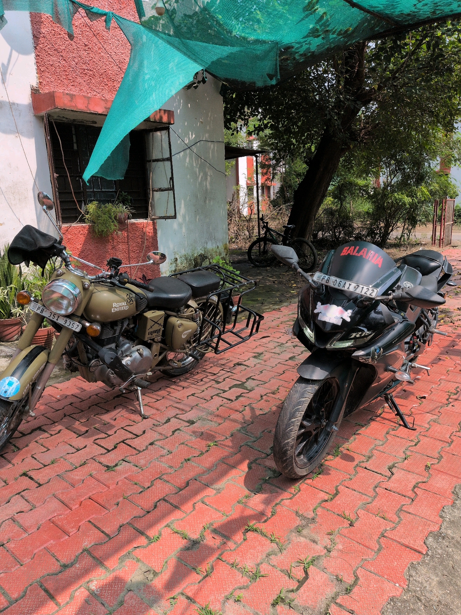 The image shows two motorcycles parked on a red brick-paved surface in front of a building. The building has a textured pink wall and a white section with a window. A green net is stretched over the area, providing shade. A large tree with dense foliage is visible behind the motorcycles. To the left, a Royal Enfield motorcycle is parked, distinguished by its olive green color and round headlight. The license plate reads "PB-35X 9357." To the right, a black sportbike is parked, featuring a red "SALARIA" sticker on its windscreen and a license plate reading "PB-06AX 7122." A bicycle is leaning against the building in the background. The scene appears to be an outdoor area, possibly a courtyard or parking space, in Pathankot, India.