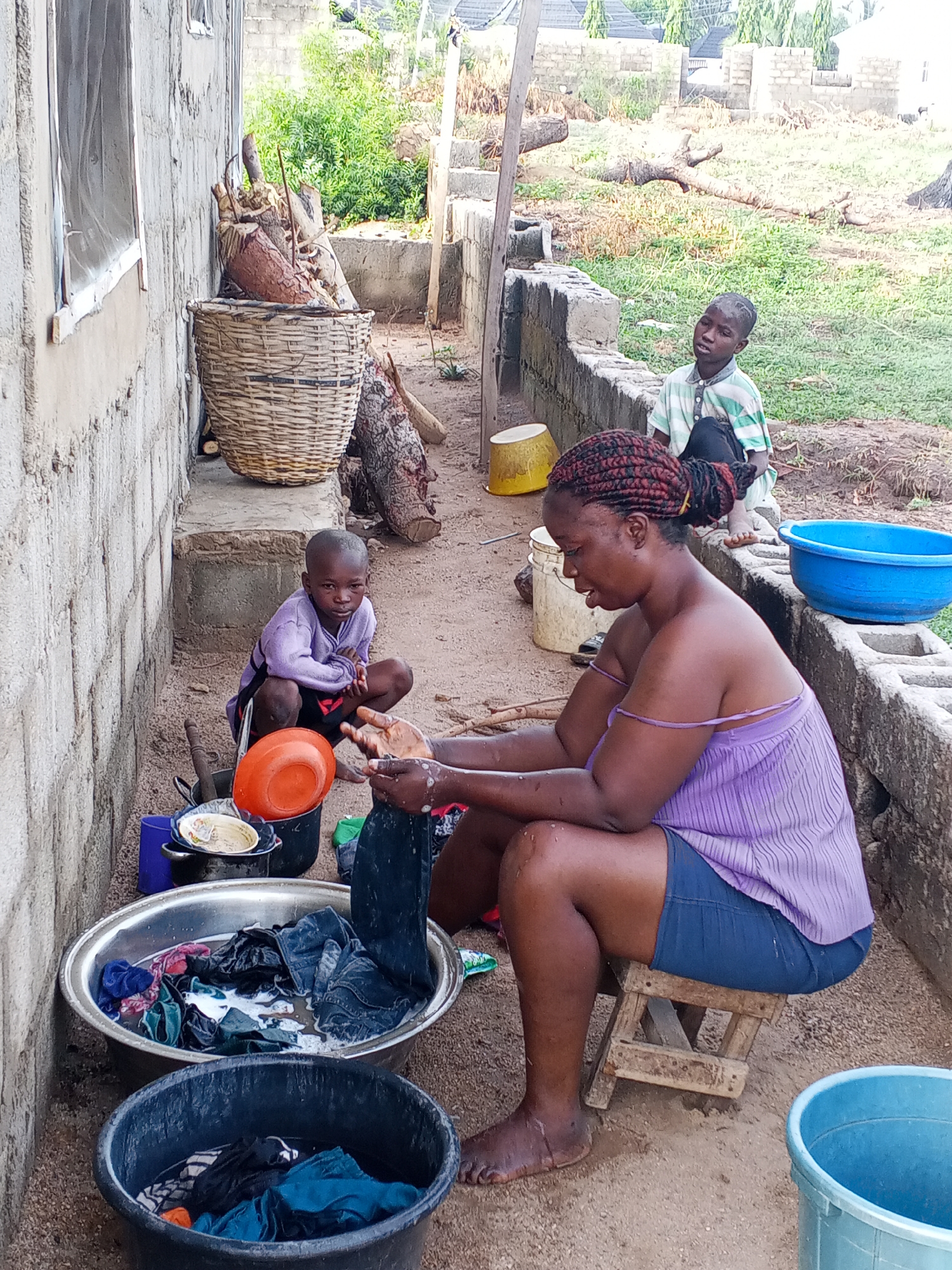 This image depicts an outdoor domestic scene in Adonkolo, Nigeria, where a woman is engaged in hand-washing clothes, accompanied by two young boys.
The main subject, a woman with braided red hair styled into a bun, is seated on a small wooden stool. She wears a light purple strap top and dark shorts, and is actively scrubbing clothes in a large, foamy metal basin. Her hands are submerged in the water, pulling out a dark garment. Beside her, on the ground, is a black basin also filled with water and clothes, suggesting multiple stages of washing.
To her left, a young boy in a purple long-sleeved shirt and dark shorts crouches on the dirt ground, intently looking towards the viewer. Further back and to the right, another boy, dressed in a green and white striped shirt, sits on a low concrete wall, observing the scene.
The setting is an unpaved yard beside a building constructed with concrete blocks. A window is visible on the wall of this building. Various household items are scattered around: a large woven basket filled with logs or firewood sits on a raised concrete platform, with more logs on the ground nearby. Several plastic buckets and basins in blue, yellow, and orange are visible, indicative of the washing process. In the background, beyond low concrete walls, lies an open area with green vegetation and some dried trees, suggesting a rural or semi-urban environment.
The image is captured in bright, natural daylight, indicating a clear day. There is no visible text in the photograph. The scene portrays a routine daily chore within a family setting.