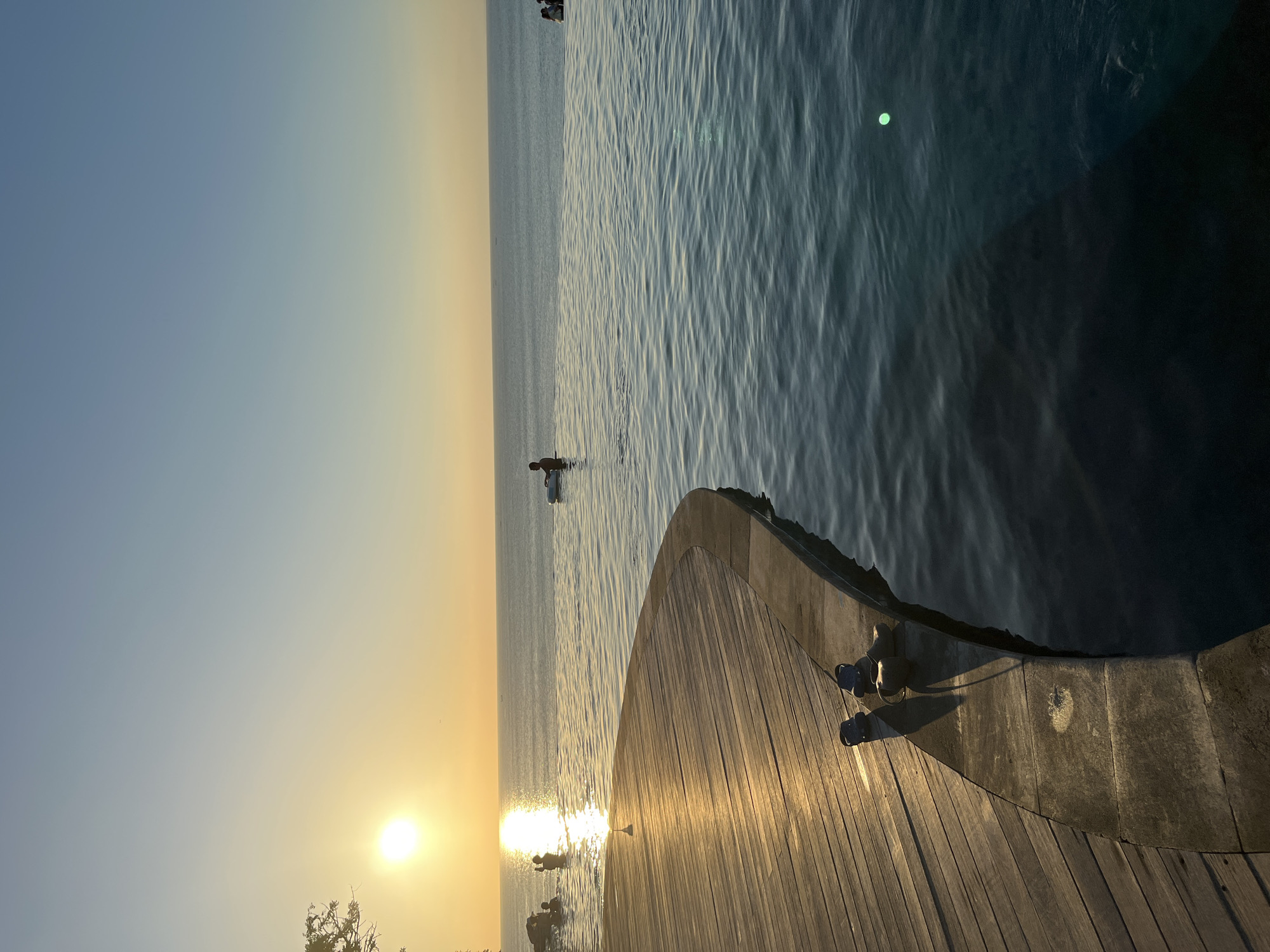 An infinity pool overlooks the ocean at sunset, casting a warm golden glow on the wooden deck. Several people are visible in the pool and the ocean. A group of individuals are swimming or wading in the left side of the pool, closer to the shore, with the sun setting behind them. Further out in the pool, a person is sitting on an inflatable tube. On the far right of the frame, a few more people are gathered near the edge of the pool. The sun, a bright orb, is low on the horizon, reflecting a shimmering path across the water. The sky transitions from a pale yellow near the horizon to a soft blue overhead. On the wooden deck in the foreground, a pair of blue clogs rest on the edge of the pool. The scene appears to be in Kuta Selatan, Indonesia, a popular tourist destination known for its beautiful beaches and resorts.