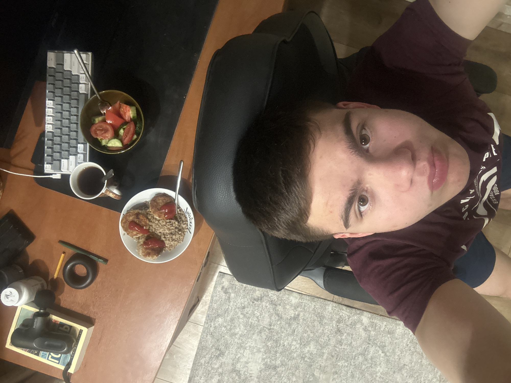 A top-down selfie shows a young man with short brown hair and brown eyes, wearing a burgundy t-shirt and blue shorts, sitting in a black gaming chair. To his left is a desk with a black mouse pad, a 60% keyboard, a bowl of salad with tomatoes and cucumbers, a mug of dark liquid, a bowl of buckwheat with meatballs and ketchup, a black ring, two writing utensils, a small white bottle, and a massager in a box. The floor is covered in light gray carpet. The location is unknown.