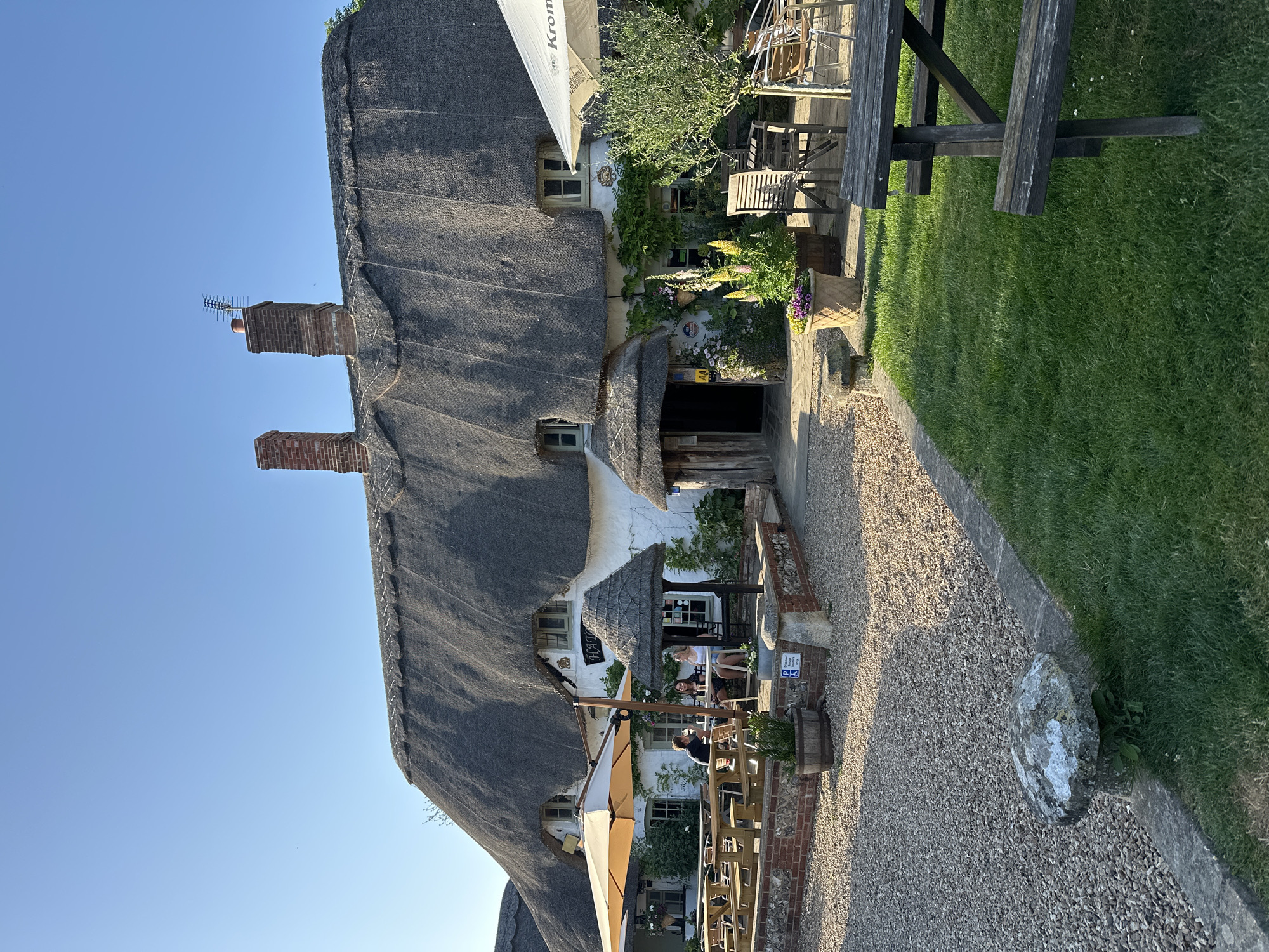 The image depicts a quaint, thatched-roof building with a traditional aesthetic. The structure features two brick chimneys rising from the roof, one of which has an antenna attached. The exterior is adorned with greenery, including vines and flowering plants, adding to its charm. Outdoor seating is arranged on a gravel patio, with tables and chairs visible. The area appears to be a pub or restaurant, as suggested by the presence of patrons seated and enjoying the ambiance. A large rock rests on the edge of the gravel path. The location is identified as being in the city of Unknown, Unknown.