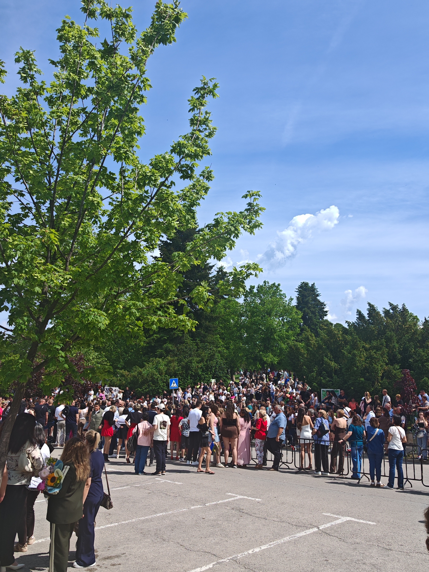 The image shows a large crowd of people gathered on a paved area, likely a parking lot, in Shumen, Bulgaria. The crowd is dense and extends towards a slightly elevated, grassy area with trees in the background. A pedestrian crossing sign is visible among the trees. In the foreground, there are people standing behind a black metal fence-like structure. Many individuals are dressed in casual attire, and some are holding flowers. The scene is outdoors on a sunny day with a blue sky and some scattered clouds.
