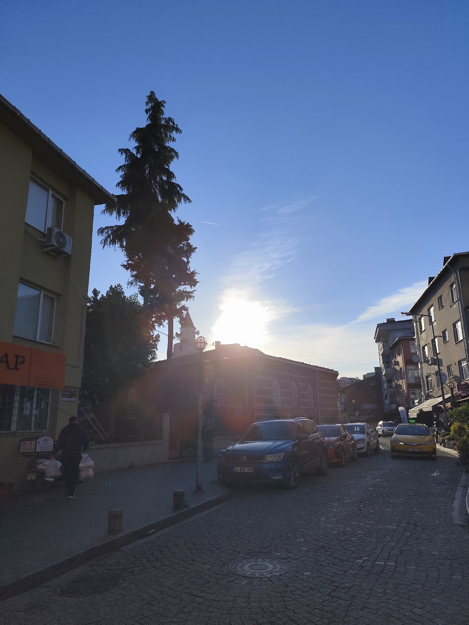 The photograph depicts a street scene in Sarıyer, Turkey. The street is paved with cobblestones and lined with parked cars, including a blue SUV with the license plate "34 MFB 100." A pedestrian, dressed in dark clothing and carrying bags, is walking on the left side of the street. On the left, there is a building with an orange sign reading "AP" and an air conditioning unit on its facade. A tall evergreen tree and other trees are visible behind the building. In the distance, a mosque with a minaret is present, obscured by the sun's glare. The sky is blue with wisps of clouds. Buildings of varying heights are on the right side of the street.