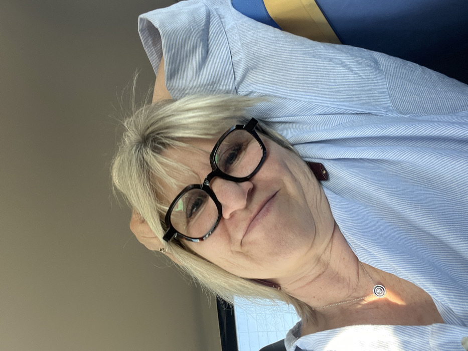 A blonde female is depicted in a close-up, selfie-style photograph taken inside what appears to be a vehicle. She wears dark-rimmed eyeglasses and a light blue, vertically striped collared shirt. A silver-colored chain necklace with a circular, possibly jeweled pendant, is visible on her chest. A dark, rectangular earring is present on her left ear. Her head is tilted, and her right arm is raised, suggesting she is resting her head on her arm or shoulder. Her expression shows a slight, closed-mouth smile. The background is blurred, indicating an indoor setting, likely a car interior, with a bright light source, consistent with daylight, entering from the left. A glimpse of what appears to be a blue and yellow striped object is visible behind her right shoulder. The specific city location of Étoile-sur-Rhône, France, cannot be confirmed from the visual information presented in the image.