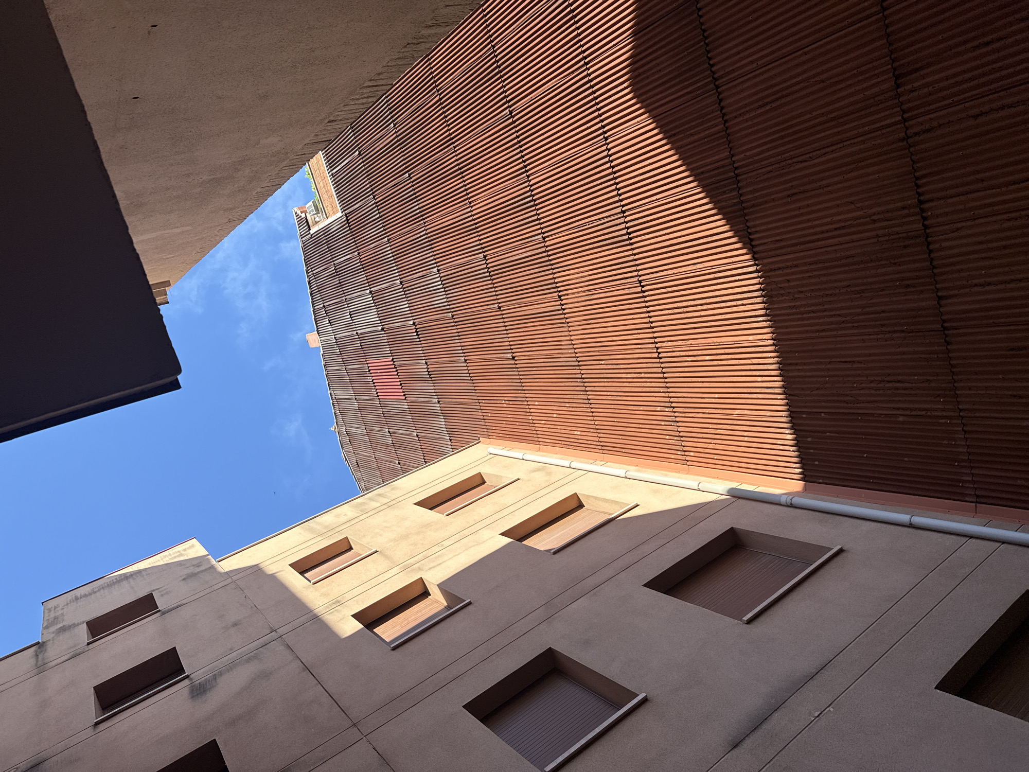 The image depicts an upward-angled architectural shot of buildings in Barcelona, Spain. The perspective is from a courtyard or alley, looking up at the sky visible between the structures. The buildings exhibit a mix of architectural styles. One building is constructed with horizontal, terracotta-colored slats and is partially covered by a shadow. Another building has a beige-colored facade with square windows, some of which have recessed window coverings. The sky is blue with few clouds. The structures are made of concrete and exhibit some aging.