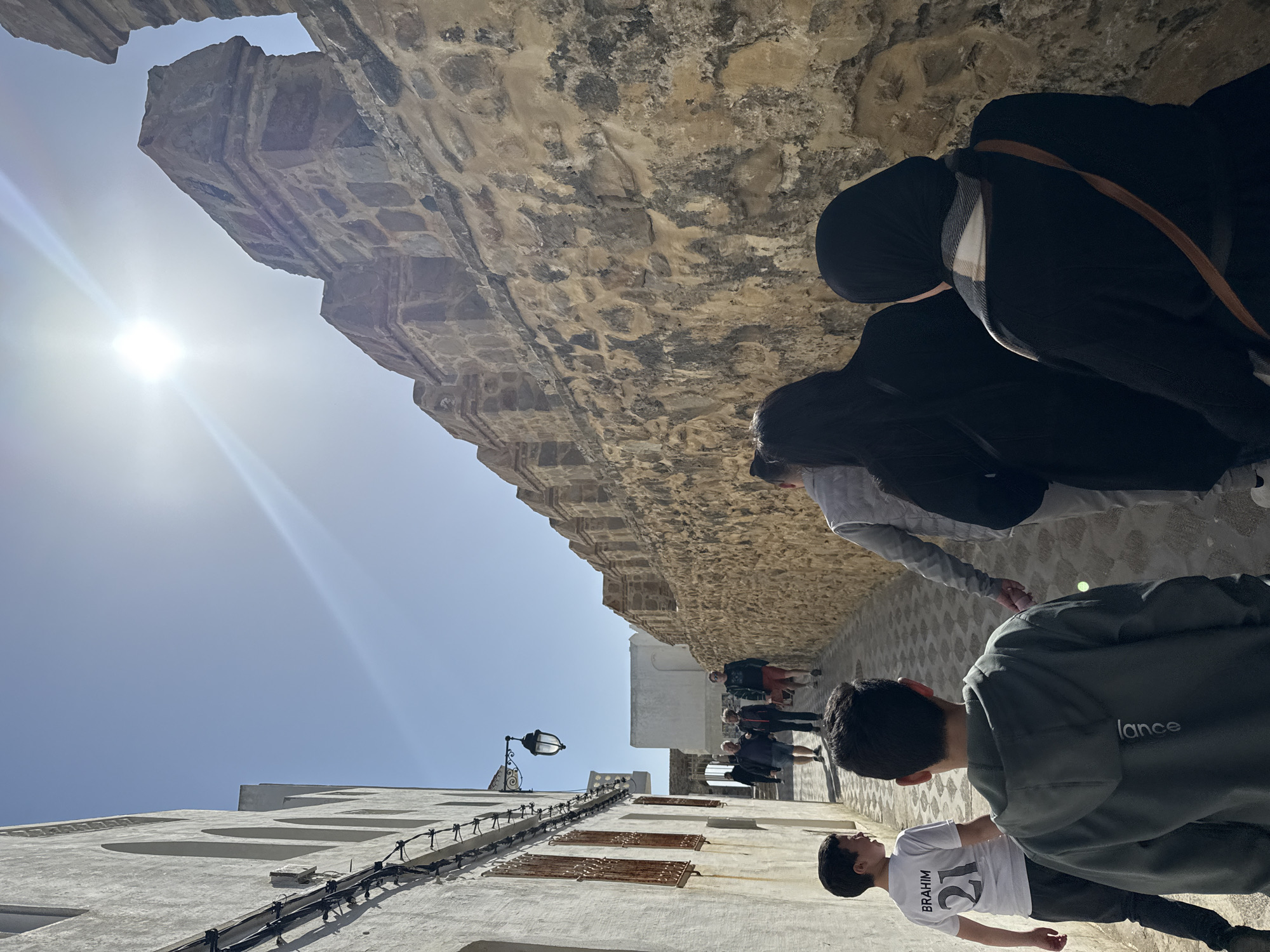 A group of people are walking through a narrow street in the city of Asilah, Morocco. The street is lined with white buildings on one side and a tall stone wall on the other. The wall appears to be part of a larger structure, possibly a fortress or ancient city wall. The sun is shining brightly, creating a glare in the sky. The people in the group are dressed in casual clothing, and some are wearing headscarves. The ground is paved with light-colored stones. A black lamppost is visible on the wall. One person in the group is wearing a t-shirt with the name "BRAHIM" and the number 21. Another person is wearing a green jacket with the word "lance" on the back. The group seems to be engaged in a walking tour or simply exploring the area.