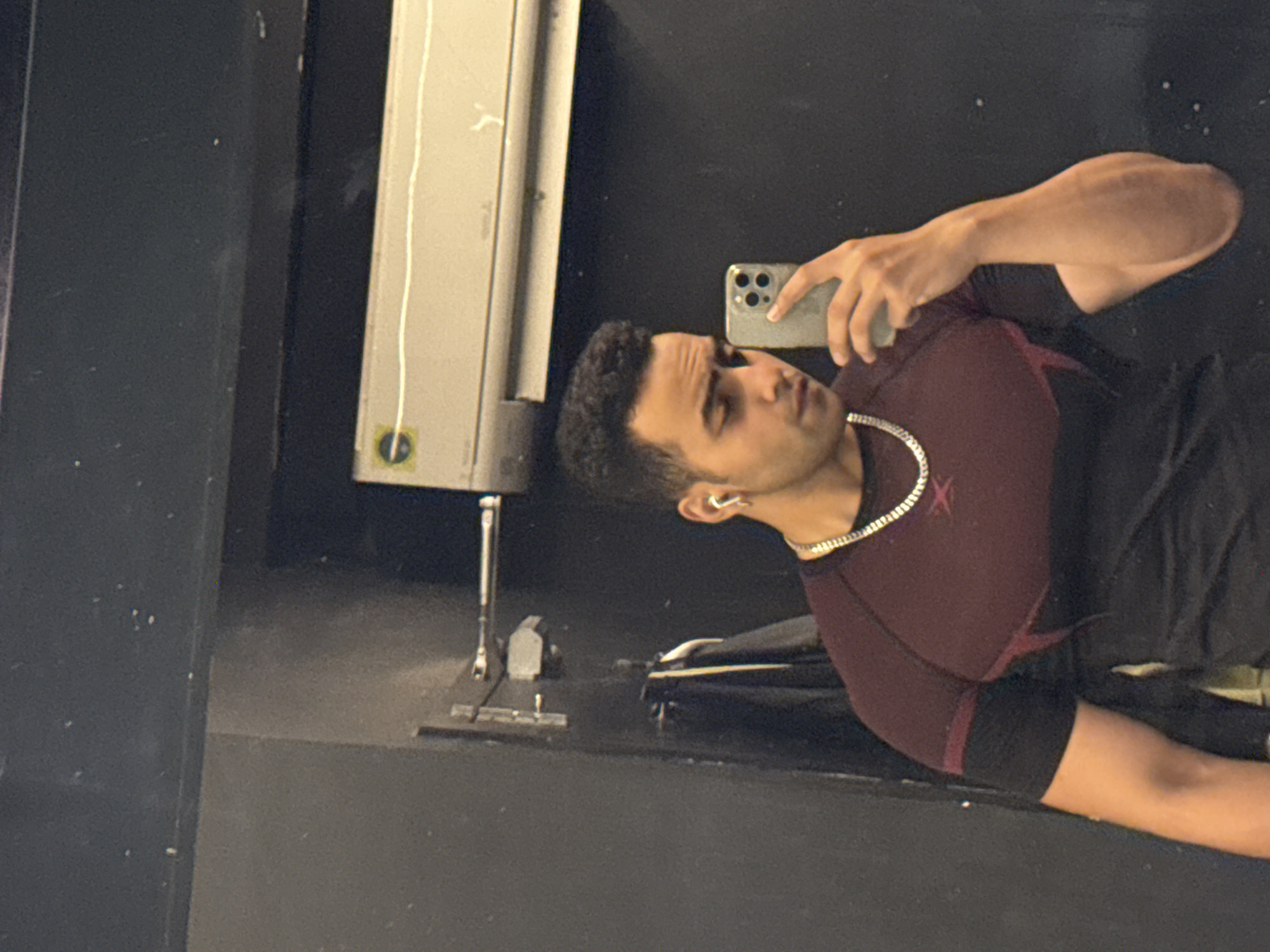 A man is taking a selfie in a mirror, possibly at a gym. The man has short dark hair, a silver chain necklace, and is wearing a burgundy and black workout shirt. He is holding a smartphone with multiple camera lenses. The background is dark, with a piece of gym equipment visible to the left. There is nothing in the image to indicate that it is in Kolkata, India.