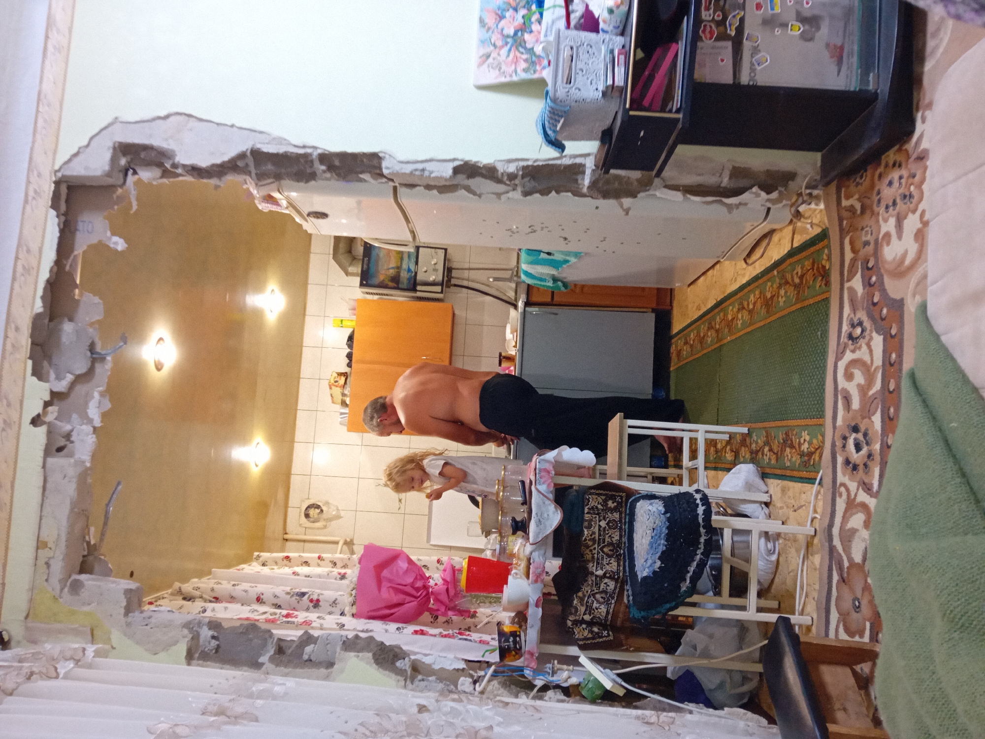 This media file captures an indoor scene in Chuhuiv, Ukraine, depicting the aftermath of some form of demolition or renovation. A large opening has been made in a wall, revealing exposed interior structures and debris.
The primary subjects are two people: an adult male, shirtless and wearing dark trousers, and a young child with blonde hair, wearing a white dress. The man appears to be performing some sort of physical activity, possibly push-ups or a plank, in the doorway of the partially demolished space. The child is standing on the other side of the opening, looking towards the man.
The setting appears to be a domestic interior, with elements of a kitchen visible, including wooden cabinets, a refrigerator, and what looks like a sink area. There are also household items scattered around, such as rugs, chairs, and fabrics. The lighting suggests it might be daytime, with warm light emanating from fixtures in the interior.
Notable details include the significant damage to the wall, indicating a disruptive event has occurred. The presence of household items and the interaction between the adult and child suggest that this was a living space.
There is no visible text that can be clearly read or is significant to the scene. The overall impression is one of a paused moment during or immediately after a disruptive event within a home.