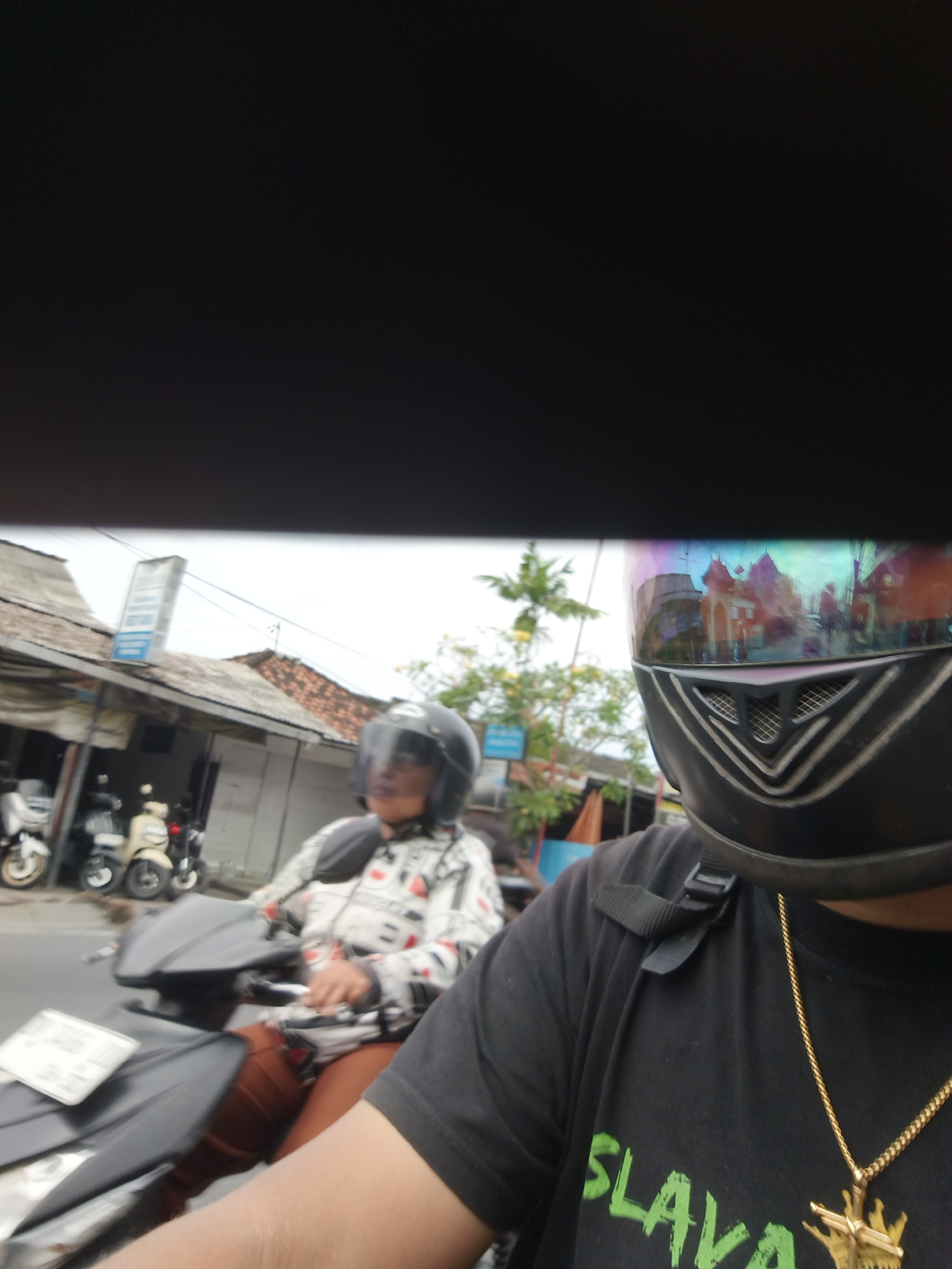 The image captures a close-up view of two individuals on a motorcycle, with the foreground dominated by the helmet of the person in the driver's seat. The helmet, black with a reflective visor showing distorted images of buildings and foliage, partially obscures the driver's face. The driver wears a black t-shirt with a gold chain and a pendant. Behind them, a passenger in a white jacket with black and red patterns is visible. The motorcycle is dark-colored. In the background, several other motorcycles are parked. The environment appears to be a street in Denpasar, Indonesia, with buildings featuring tiled roofs and what looks like a street sign. The lighting suggests it is daytime, possibly overcast.