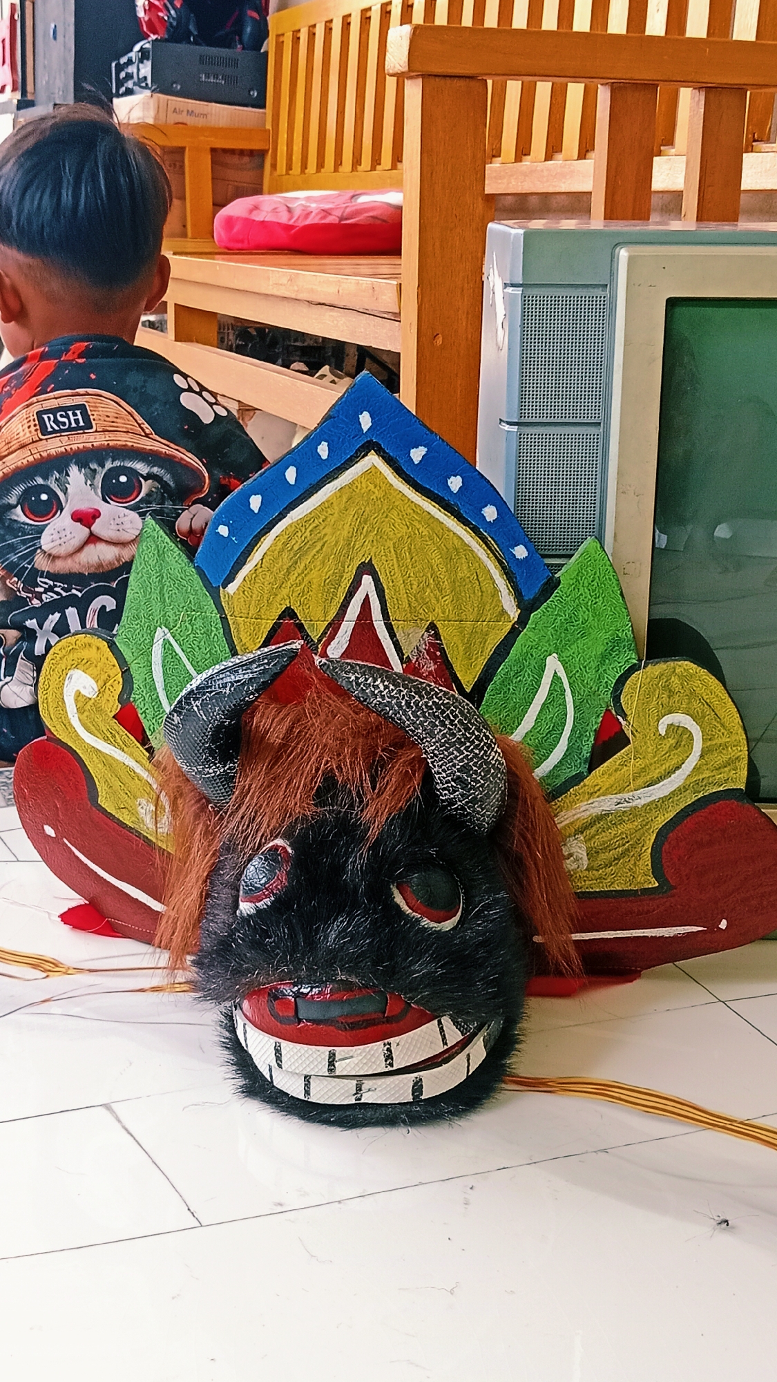In the city of Karang Dukuh, Indonesia, a close-up, eye-level shot captures a partially visible young boy from behind, facing away from the camera. He is wearing a black t-shirt adorned with a cartoon cat wearing a straw hat, an image further detailed by the letters "RSH" on the hat. The boy is seated in front of a wooden bench, with a portion of a wooden structure visible behind him. To the right of the bench, a vintage television set with a light blue casing and a dark screen stands. In the foreground, a colorful, stylized mask resembling a bull or buffalo is placed on a white tiled floor. The mask is predominantly black with red eyes and a wide, grinning mouth featuring white teeth. It is adorned with furry brown hair and two black horns with a textured pattern. The mask is decorated with vibrant painted elements in red, yellow, green, and blue, creating a festive and somewhat abstract design. The environment appears to be indoors, possibly in a home or a workshop, with natural lighting illuminating the scene. The floor is composed of white square tiles.