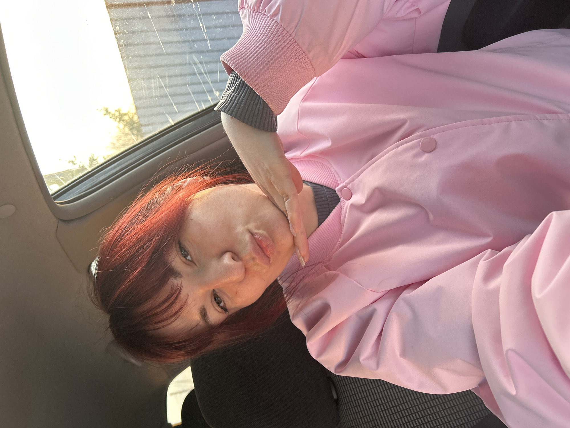 A selfie shot of a woman with red hair wearing a light pink jacket with gray sleeves. The setting appears to be inside a car, based on the visible car interior. Sunlight is filtering through the car window. The woman is holding her hand to her face in a playful pose. The specific location within the city of Фоки, Russia is not identifiable from the visual information available.