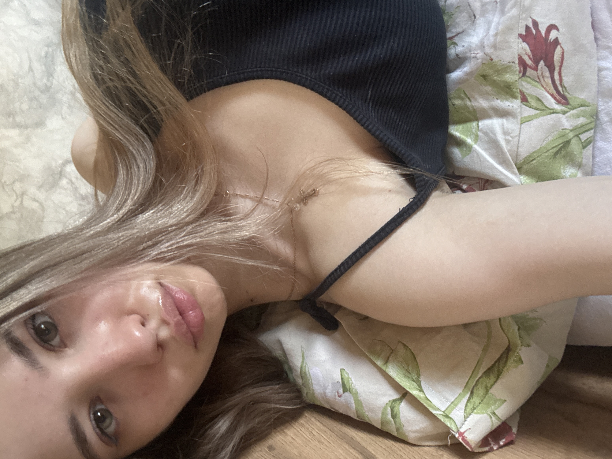 A woman with light brown hair and fair skin is lying down. She is wearing a black tank top and a gold necklace. She is resting on a pillow with a floral pattern, which is set on a wooden surface. The location is not discernible.