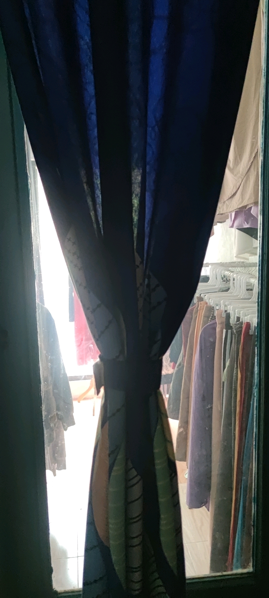 The image depicts a scene viewed through a window or doorway, partially obscured by a dark blue curtain with a patterned tie-back. To the left, a dark green frame is visible, possibly a door or window frame. Beyond the curtain, the scene appears to be an indoor space with clothing hanging on a rack. The clothing items are varied in color, including shades of purple, brown, red, and yellow. Some items are on hangers, suggesting they are being stored or displayed. The lighting suggests it may be daytime, with light filtering in from the outside. The specific location in Gambarsari, Indonesia, is not determinable from the visual information alone. There are no people visible in the image.