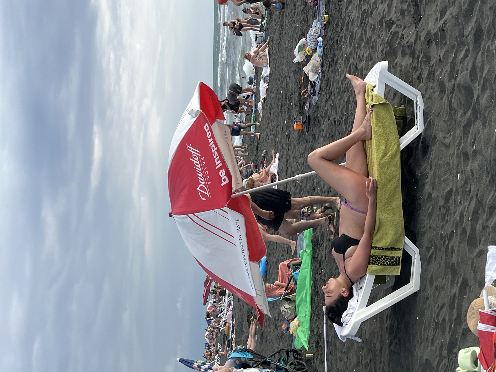 An overcast day at Ureki Beach in Georgia. The dark sand is covered with beachgoers and umbrellas. In the foreground, a woman lies on a white lounge chair, reclined on a green towel. She is wearing a black bikini and purple bottoms, with her eyes closed and mouth open as if she is speaking or yawning. A red and white umbrella, branded "Davidoff EVOLVE be inspired," is positioned behind her. Further back, other people are visible on lounge chairs, sitting on towels, and walking along the shore. The sea is visible in the background, with moderate waves.
