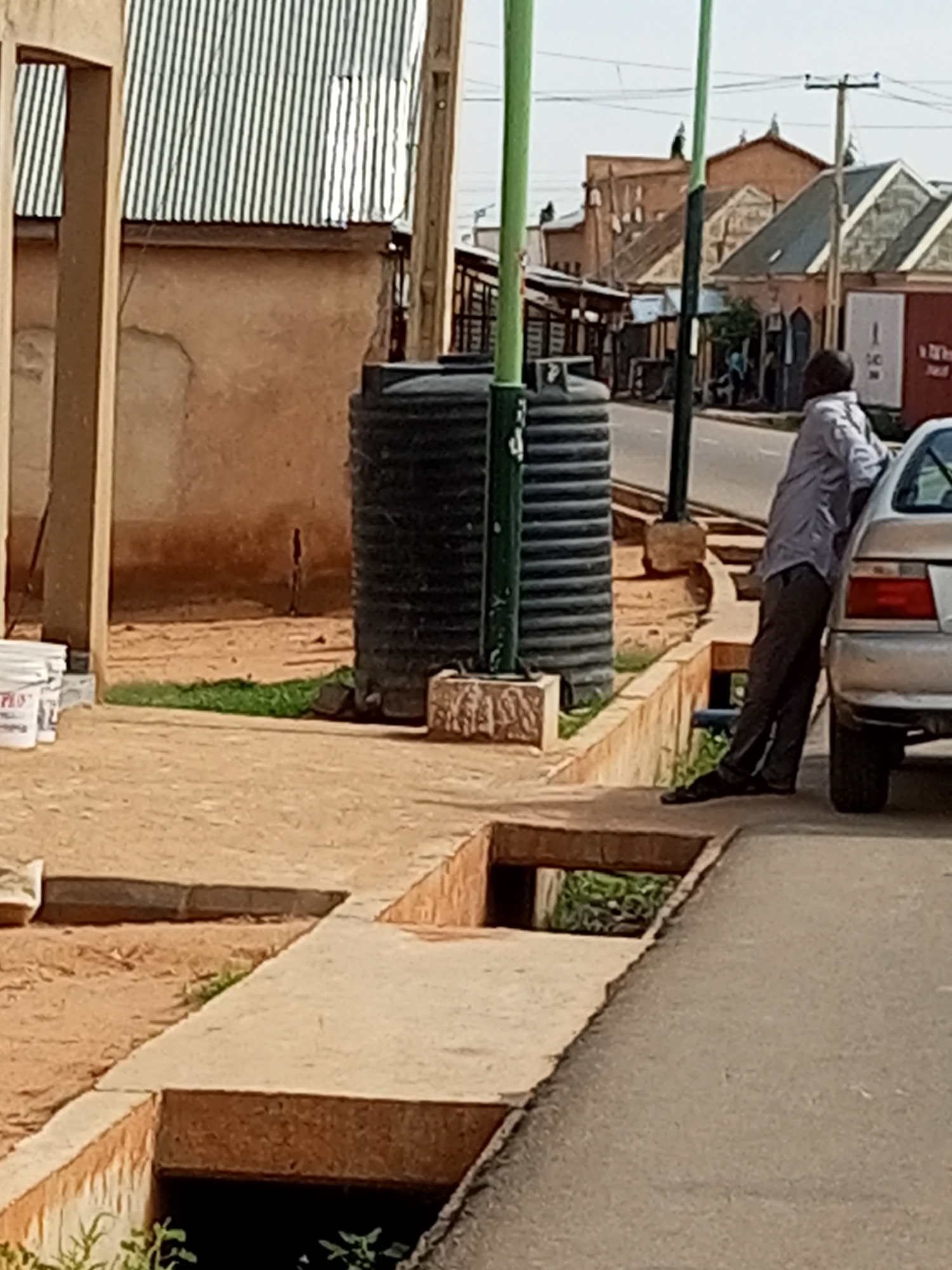 The image shows a street scene in Tempure, Nigeria. A man in a light blue shirt and dark pants is leaning against a silver car parked on the side of a road. He appears to be looking down and to his left. In the background, there is a large black water tank, a building with a corrugated metal roof, and other structures with similar roofing. A drainage ditch runs along the side of the road in the foreground. The overall environment suggests a typical urban setting in Nigeria.