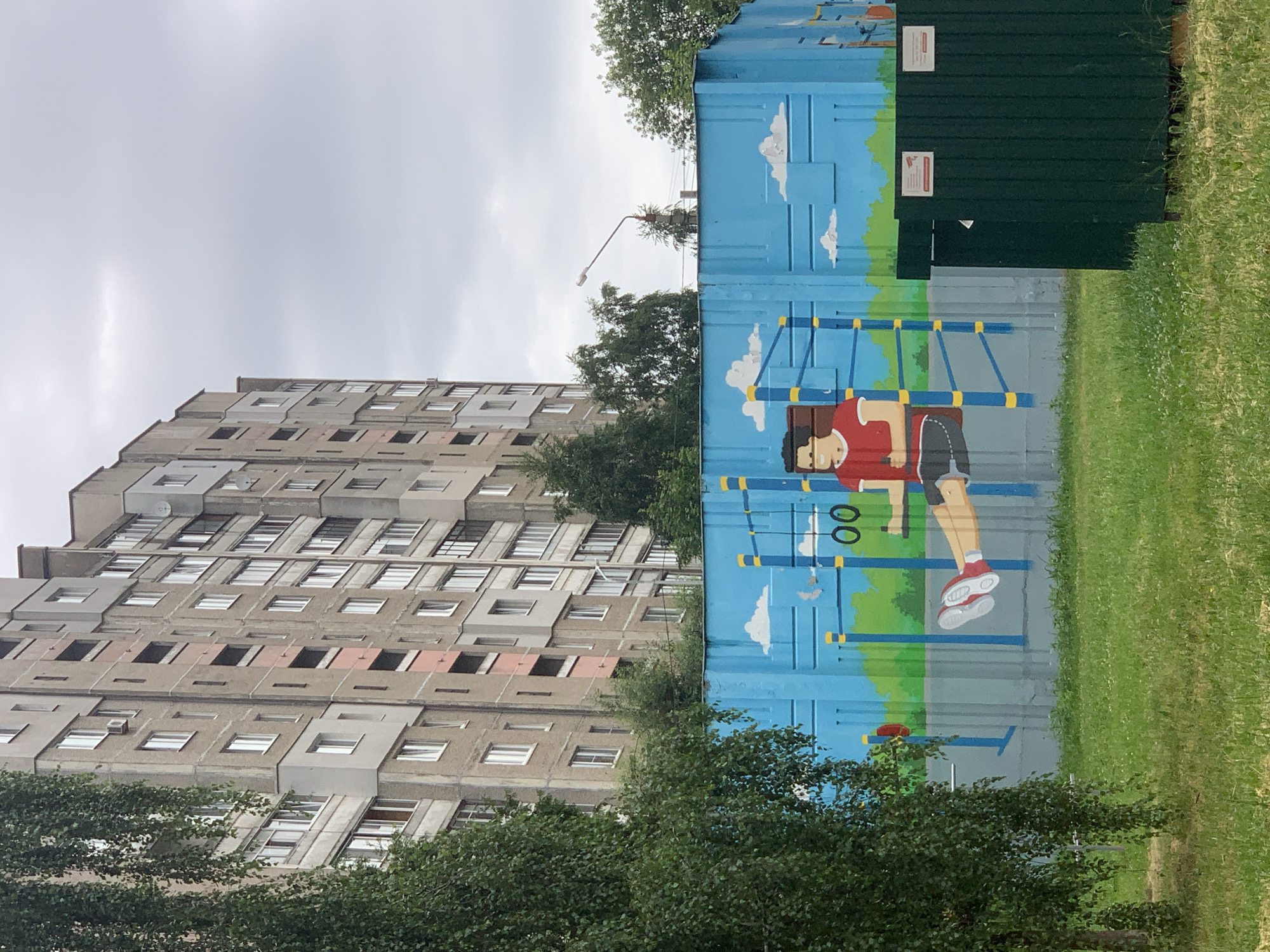 The image captures an outdoor scene in the city of Minsk, Belarus. In the foreground, a lush green lawn is visible with scattered white flowers. To the left, a large tree with drooping branches partially obscures the view. A vibrant mural adorns a blue wall, depicting a cartoonish figure performing exercises on a pull-up bar. The figure is wearing a red t-shirt and shorts, with red sneakers. The mural also features clouds and a stylized green landscape. Behind the mural, two dark green trash receptacles stand side-by-side, each with a white sign attached. To the left of the trash receptacles, another, smaller red exercise ring is painted on the mural. In the background, a tall, grey, multi-story apartment building dominates the skyline. The building has numerous windows and balconies, and its architecture suggests a Soviet-era design. The sky above is overcast and grey, indicating a cloudy day.