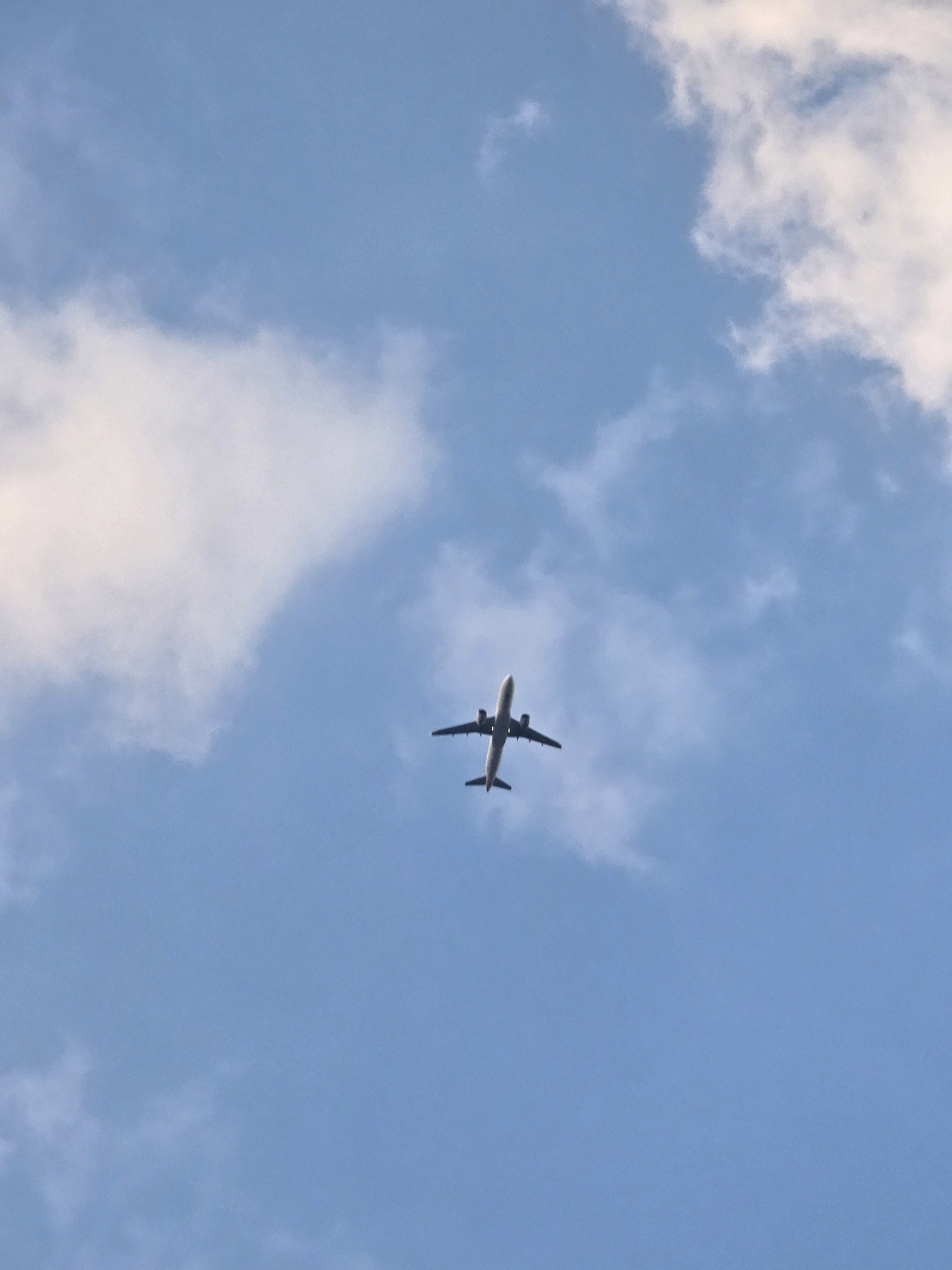 An airplane is flying in a blue sky with scattered clouds. The airplane is silhouetted against the sky and appears to be a commercial jetliner. The image was taken from below the airplane, looking up. The specific location in Podgorica, Montenegro is not discernible from the image.