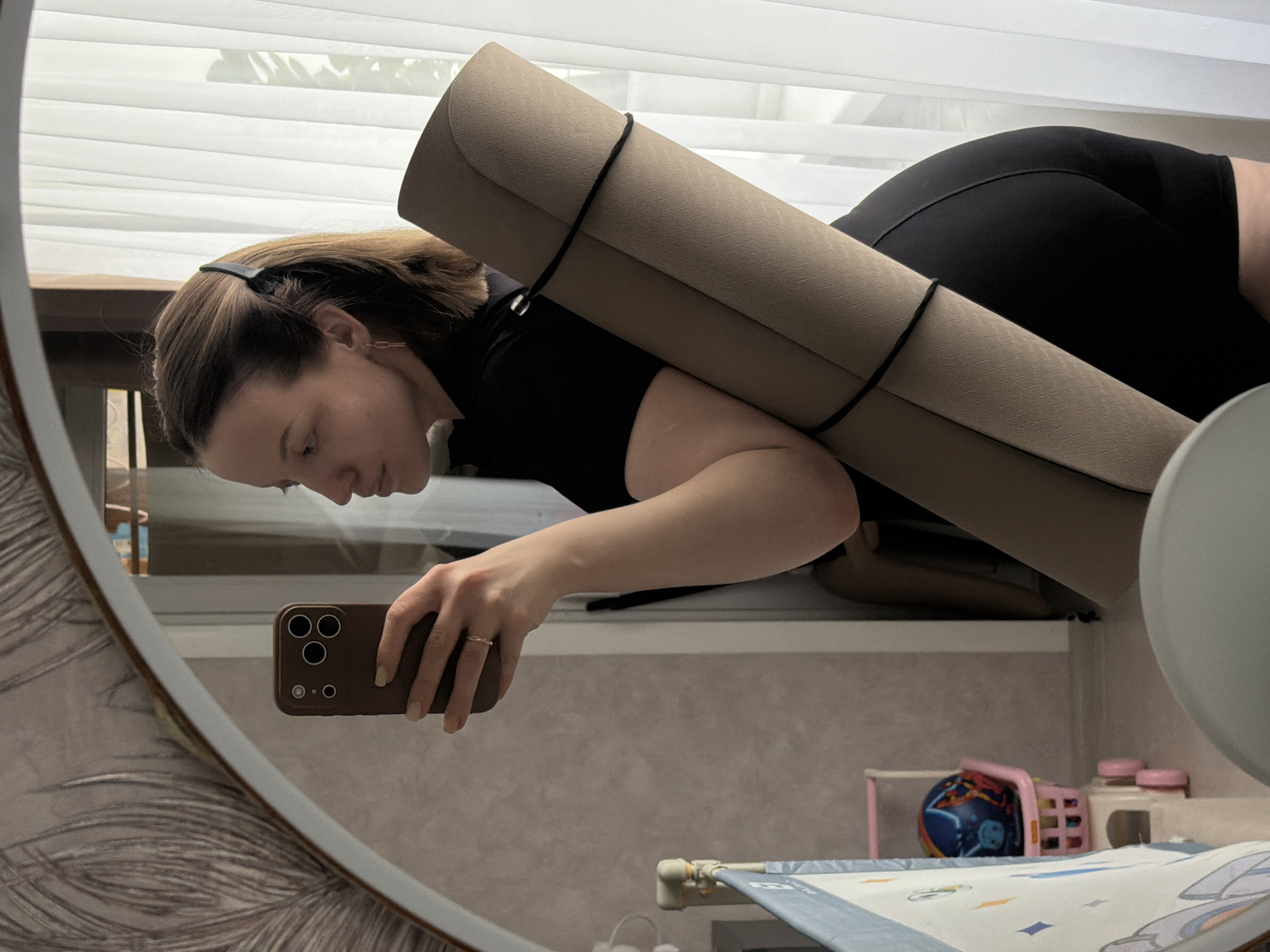 A horizontal full-shot image captures a woman's reflection in a large, round or oval mirror in an indoor setting. The woman is positioned horizontally across the frame, seemingly lying on her stomach or leaning heavily forward/downward. She has shoulder-length brown hair, pulled back with a black headband, and is wearing a black top and black shorts or leggings. Her left hand holds a brown smartphone, equipped with a three-lens camera system, as she takes a mirror selfie. A light-colored ring is visible on her left ring finger.
A large, light brown rolled mat, secured with black straps, is resting across her back and shoulders, suggesting preparation for or completion of an activity like yoga or exercise. In the background of the reflection, white horizontal blinds are visible through a window, indicating daytime. Below the window, there's a light-colored shelf or surface. To the lower right, within the mirror's reflection, parts of a child's play area are visible, including a small pink toy goal, a blue and red ball, and other small toys resting on a white and blue patterned mat or sheet. The wall visible in the reflection has light-colored, textured wallpaper. The scene is located in Reshetnikovo, Russia. There is no visible text.