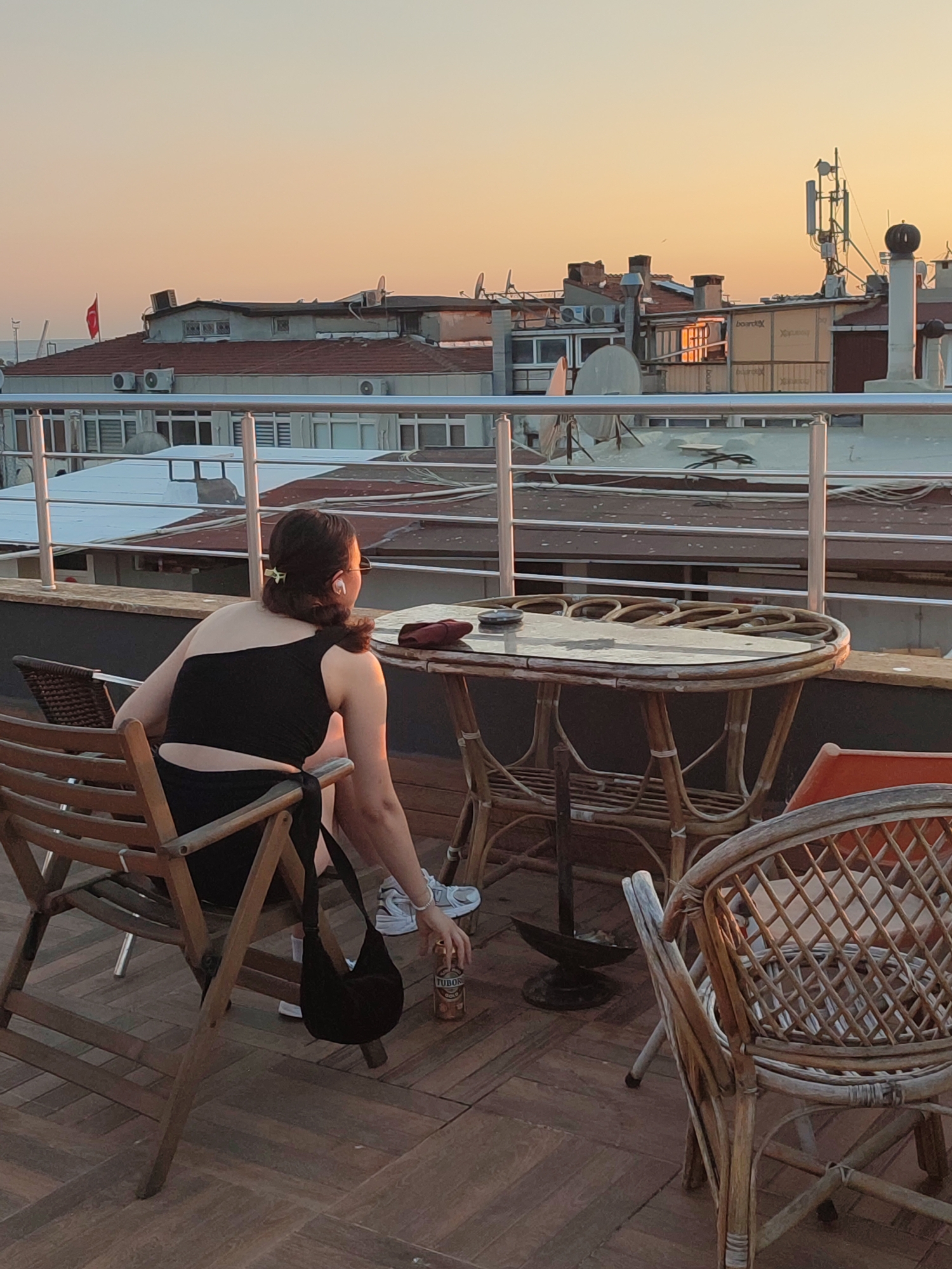 A woman sits on a wooden chair on a rooftop patio in Kadıköy, Turkey, facing away from the camera and looking out at the sunset over the city. She is wearing a black one-shoulder top and a black skirt. She has earbuds in her ears and her hair is pulled back. A black purse is hanging from her chair. On the table next to her is a can of Tuborg beer and an ashtray. The patio is furnished with several wicker chairs and a glass-topped table. In the background, buildings with red-tiled roofs and satellite dishes are visible. The sky is a gradient of orange and yellow, indicative of a sunset.