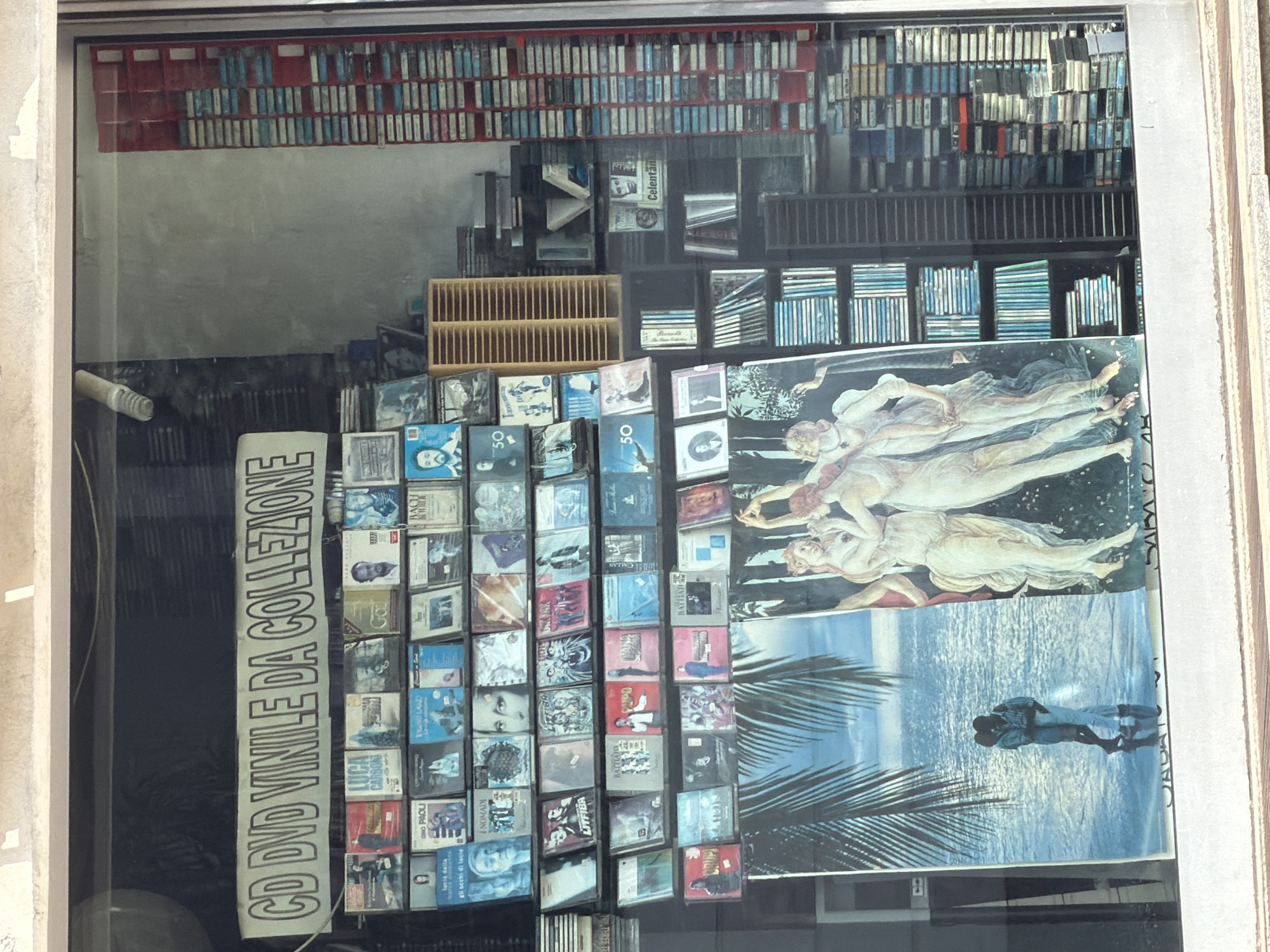 The image displays the contents of a shop window, likely a music or media store, viewed from the outside with reflections on the glass. The shop is located in Vigevano, Italy. The window is filled with a vast collection of physical media, primarily CDs, DVDs, and what appear to be cassette tapes or boxed sets, arranged on shelves and in stacks.
At the top, multiple rows of cassette tapes or small rectangular media cases are neatly organized within red and dark shelving units. Below these, numerous CD and DVD cases are displayed, some stacked horizontally and others vertically, showcasing various album covers. A prominent vertical sign in the lower left reads "CD DVD VINILE DA COLLEZIONE," translating to "CD DVD VINYL FOR COLLECTION," indicating the nature of the items.
Several Italian artists and bands are identifiable from the album covers, including LUCA CARBONI, GINO PAOLI, BAGLINI, LUCIA DALLA, I NOMADI, LITFIBA, and CELENTANO. Some covers also feature the number "50," possibly signifying special editions or anniversaries.
Beyond the media, the display incorporates large visual elements. A significant portion of the right side features a classical art reproduction, depicting several nude figures in a style reminiscent of Botticelli's "The Birth of Venus." Below this, a poster shows a couple embracing on a beach with palm fronds, overlooking sparkling water. The presence of reflections suggests it is daytime. The overall scene conveys a shop dedicated to collectible music and video media, offering a nostalgic and comprehensive selection.