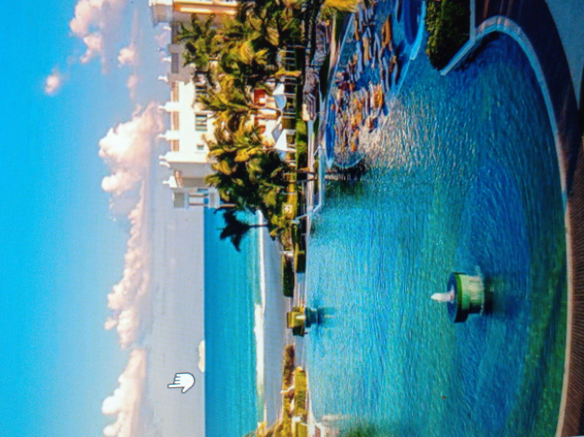 The image depicts a luxurious resort with a large, multi-level swimming pool in the foreground, which has a fountain in its center. The pool area is surrounded by lounge chairs and palm trees. In the background, a white resort building with a Mediterranean architectural style is visible, situated near the ocean. The ocean is calm with gentle waves, and the sky is a clear blue with scattered white clouds. A distant ship can be seen on the horizon. There are no identifiable landmarks or features that would place this scene within the city of Mukachevo, Ukraine. The overall impression is a tropical vacation destination.